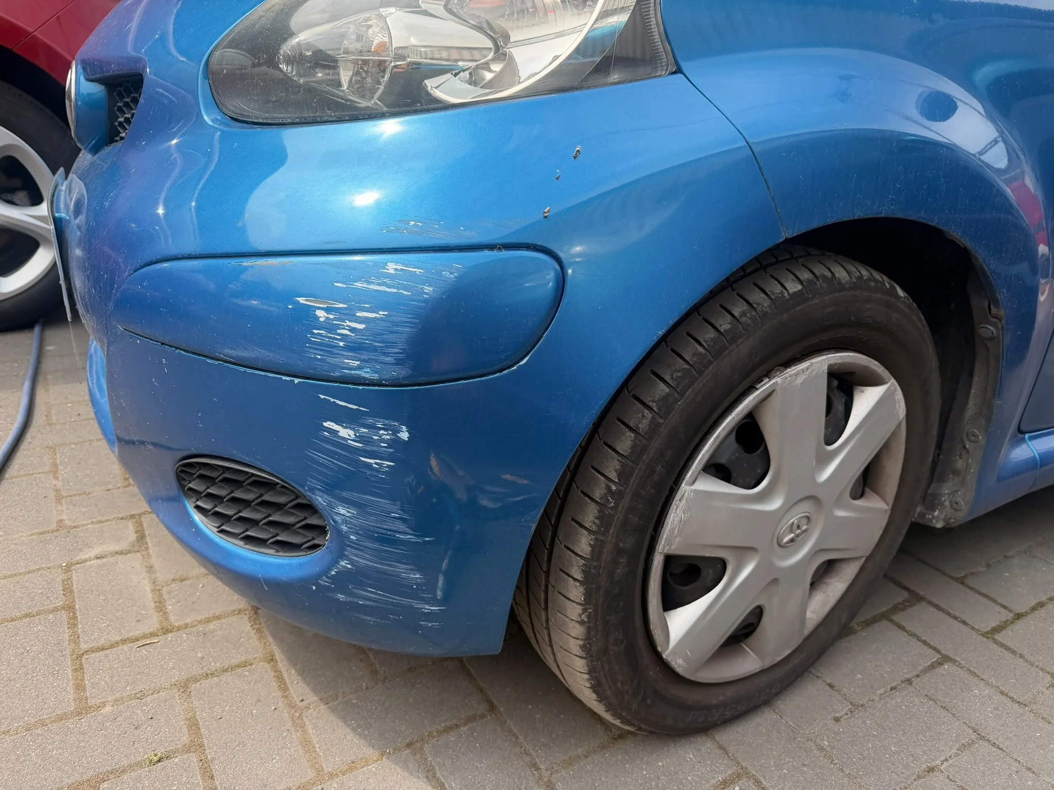 Close-up of a blue car with scratches and small dents on the front bumper and fender, parked on a paved surface.