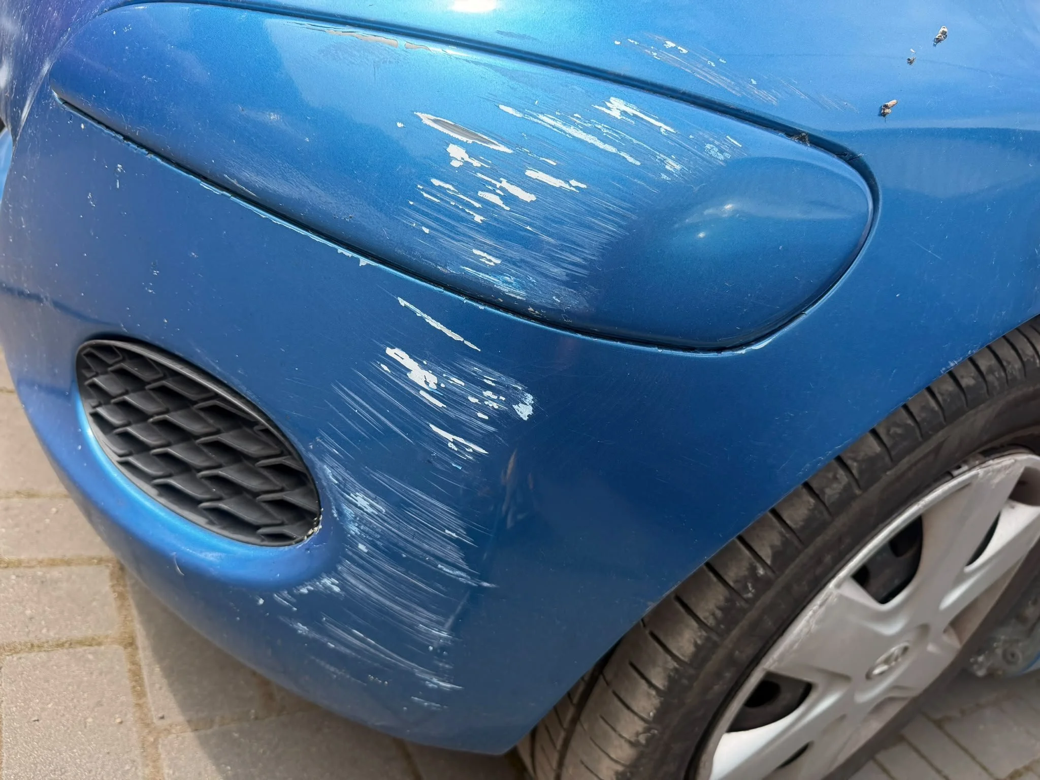 Blue car with visible scratches and paint damage on the front bumper near the left headlight.