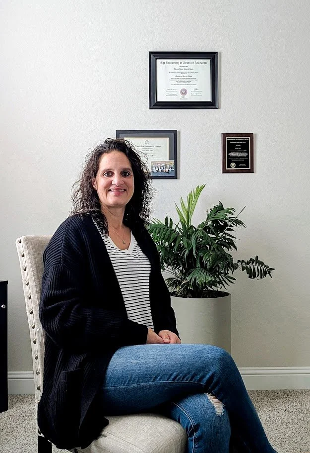 A woman with curly dark hair smiling and sitting on a beige chair in an office, with a potted plant and framed certificates on the wall behind her.