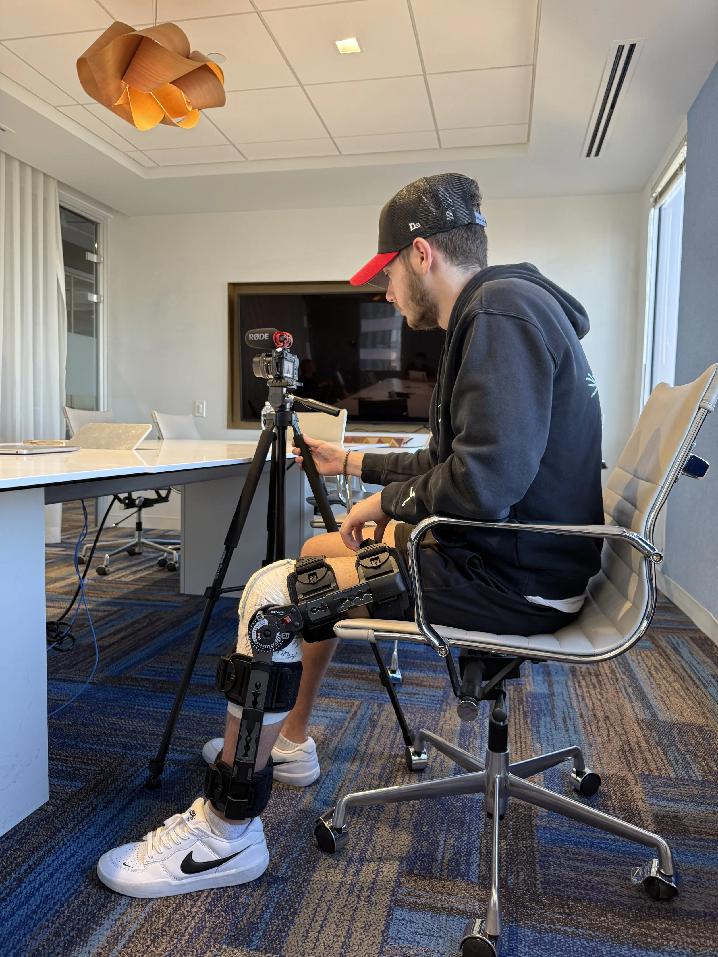 A young man with a beard, wearing a black hoodie, white sneakers, and a black and red cap, is sitting in an office with a large TV screen on the wall behind him. He is looking down at his phone, which he is holding in his right hand. He has a leg brace on his right leg and is seated on a white office chair with armrests. There is a camera on a tripod in front of him, filming or photographing him. The office has a patterned carpet, a long table with chairs, and large windows with curtains.