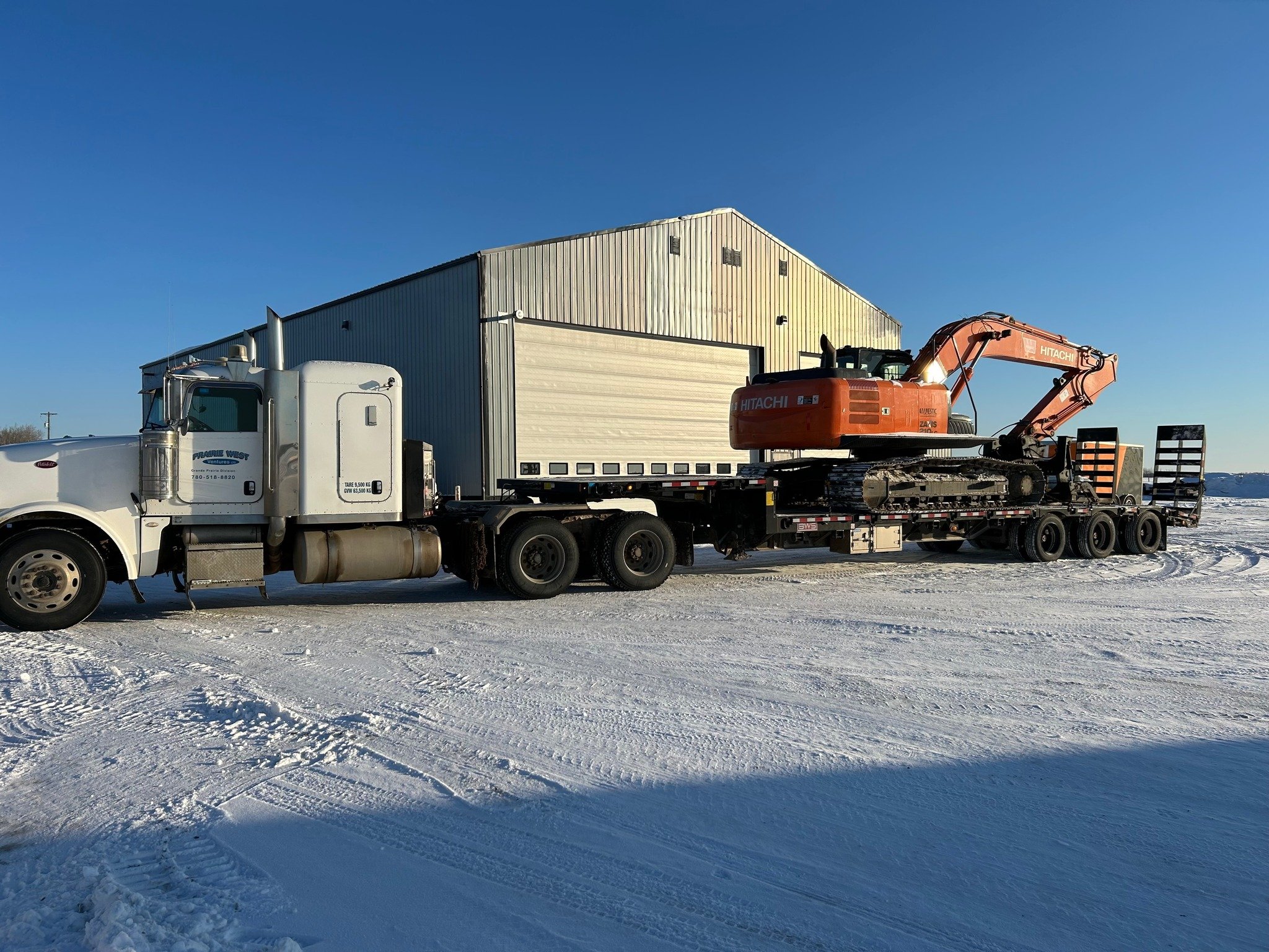 A semi-truck with a trailer carrying an orange excavator on a snowy ground under a clear blue sky.