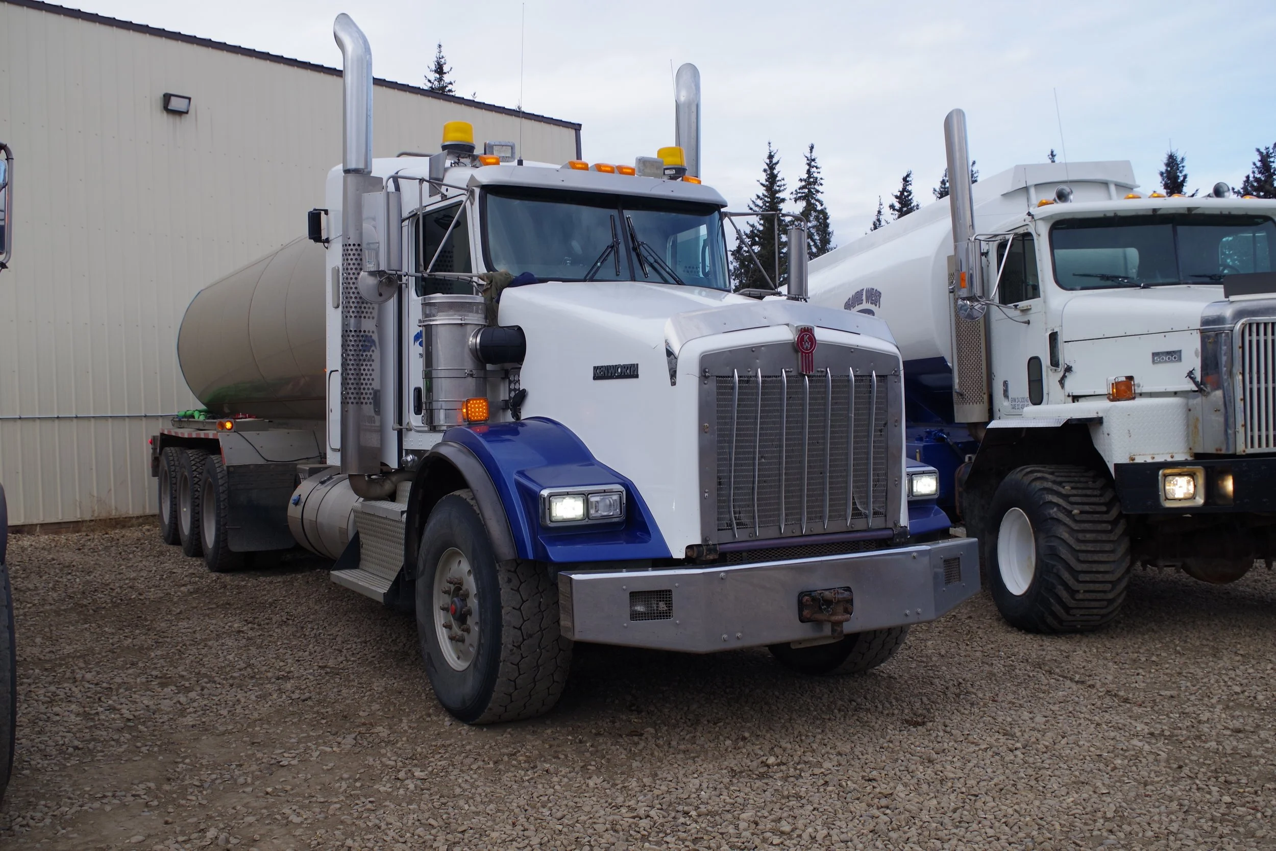 Two large tanker trucks parked outdoors in front of a beige building, with trees visible in the background.