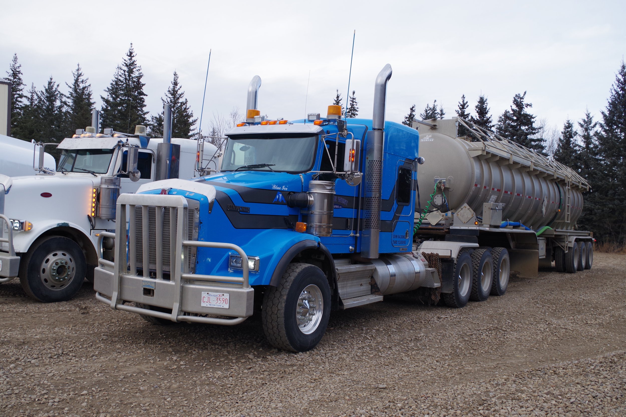 A blue semi-truck with a large cylindrical tank trailer parked on a gravel lot, with trees and a cloudy sky in the background.