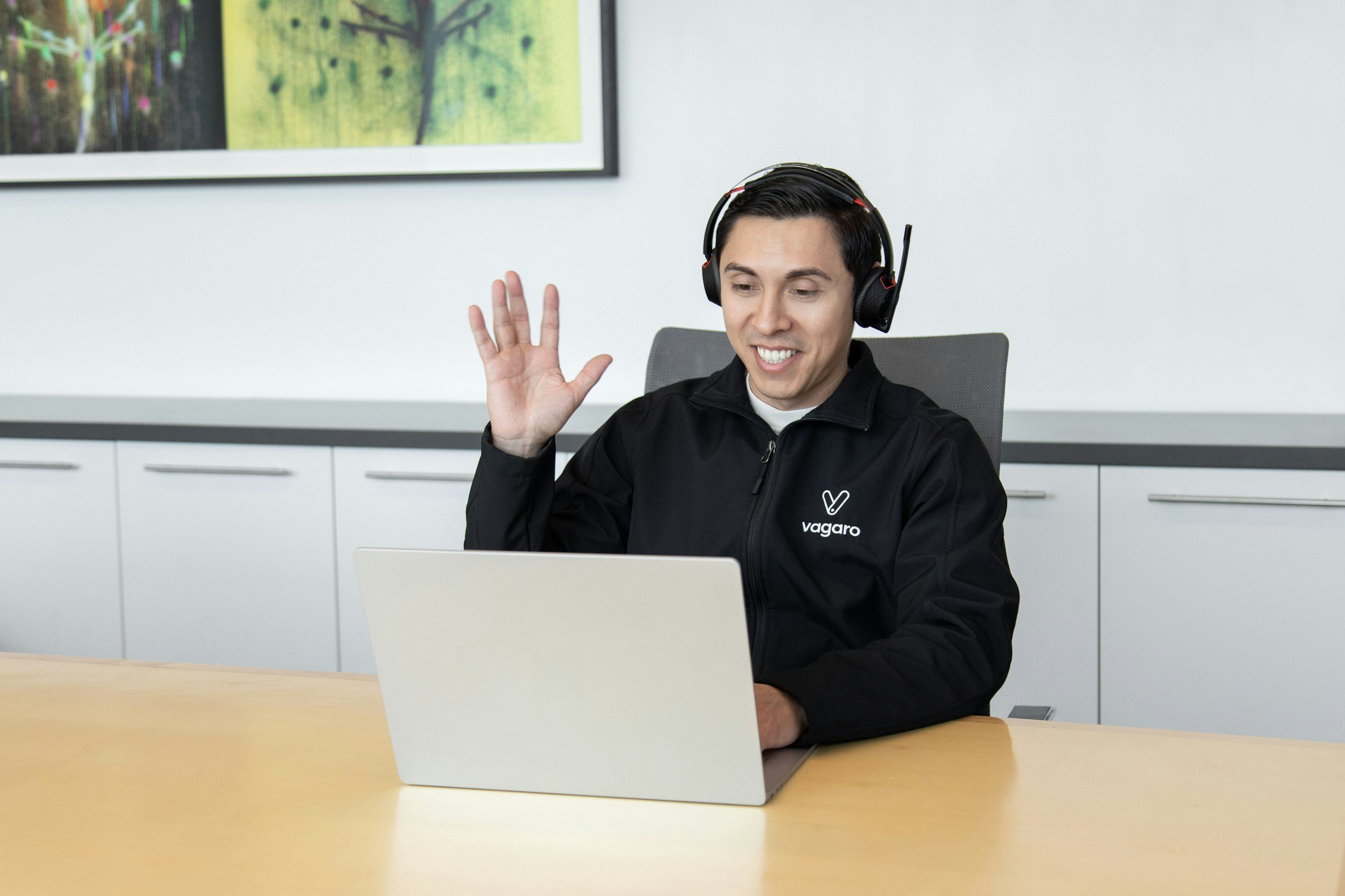 A man wearing a black jacket and headphones sitting at a desk with a laptop, smiling and waving during a video call in a modern office setting.