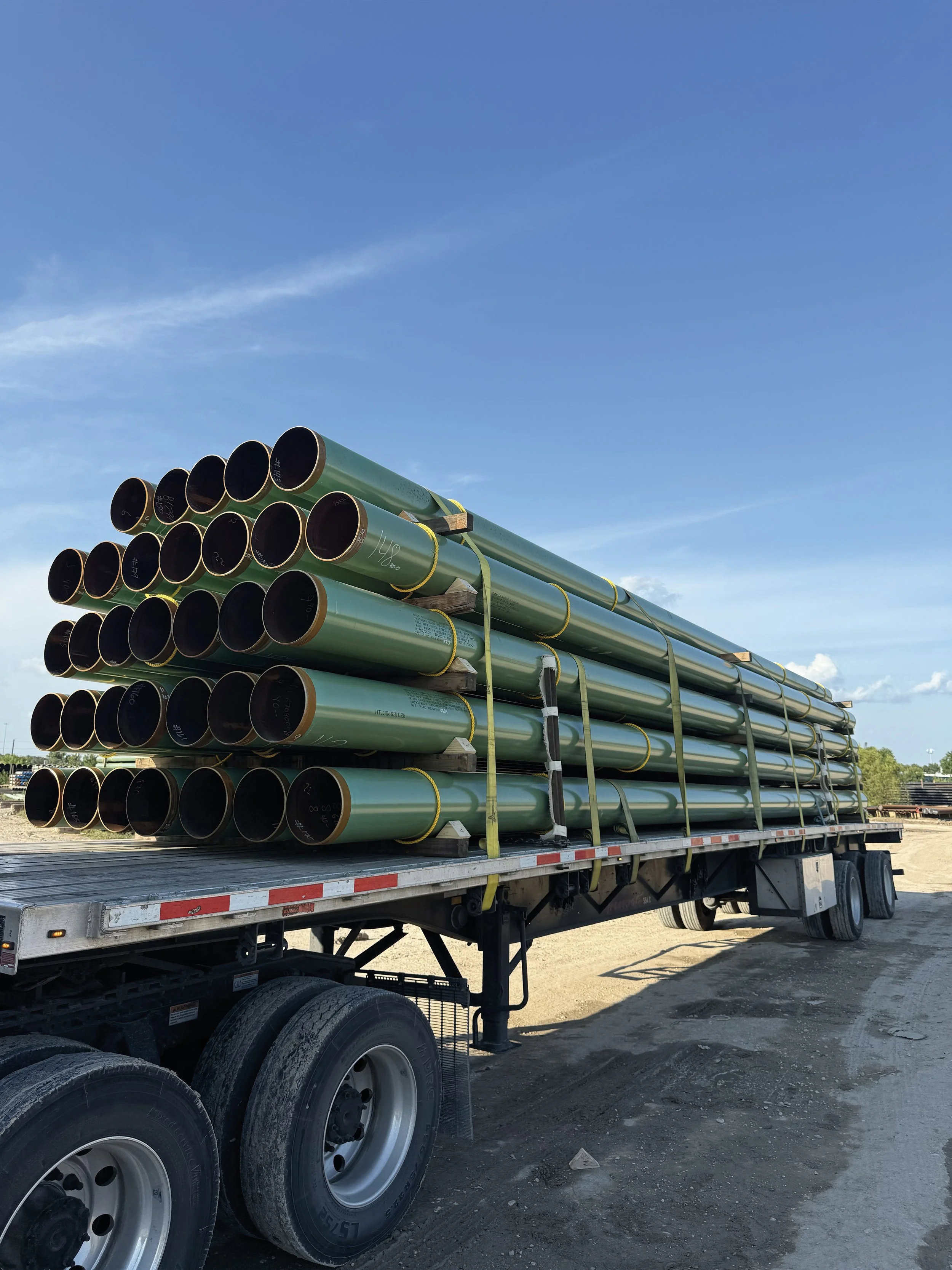 A flatbed truck transporting large green metal pipes stacked horizontally in multiple layers. The truck is parked on a dirt surface, with a clear blue sky above.