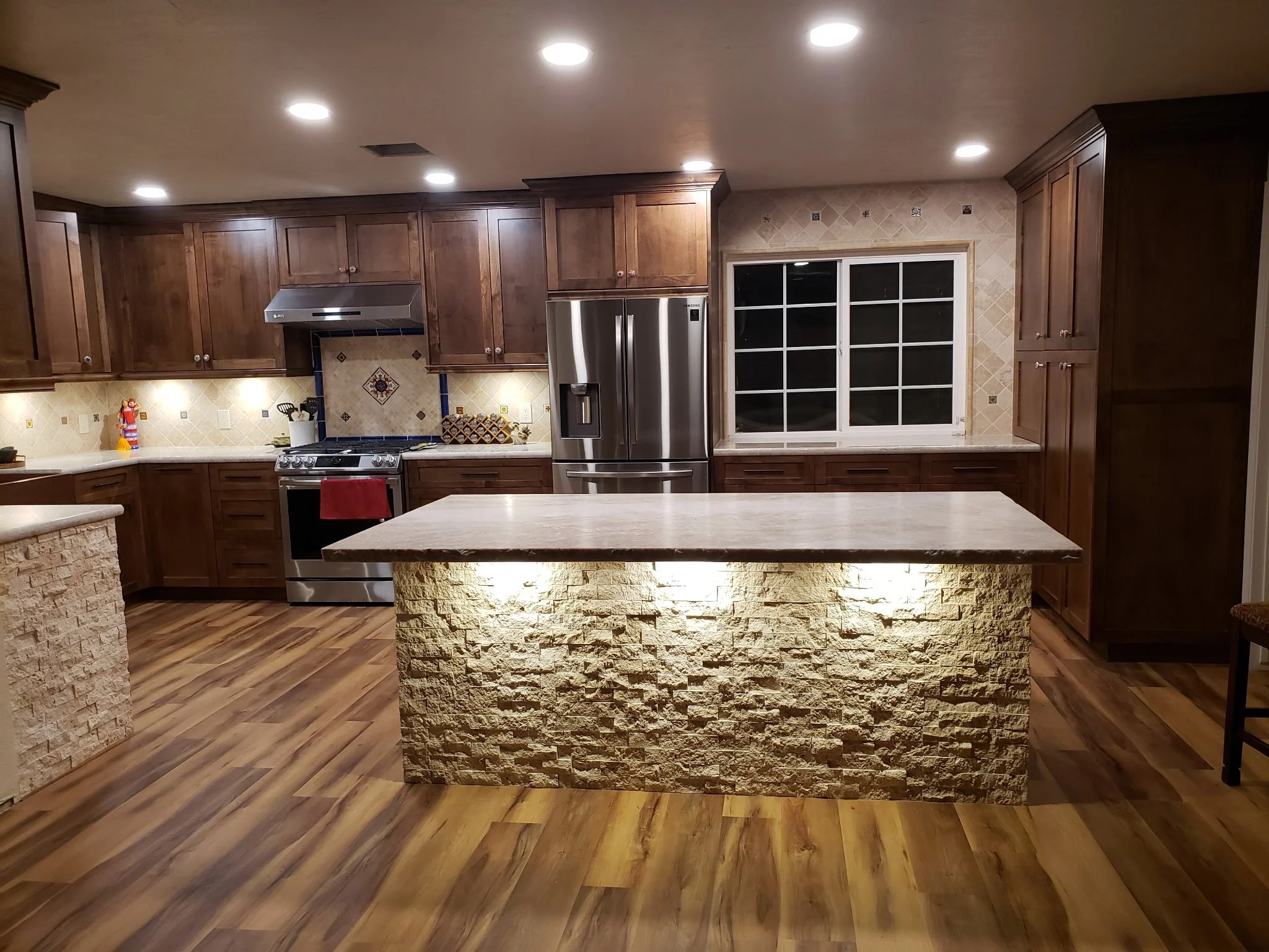 A spacious kitchen with wooden cabinets, stainless steel refrigerator, stove, and beige tiled backsplash. A stone kitchen island with a matching countertop is in the foreground, with recessed lighting on the ceiling and a window on the right wall.
