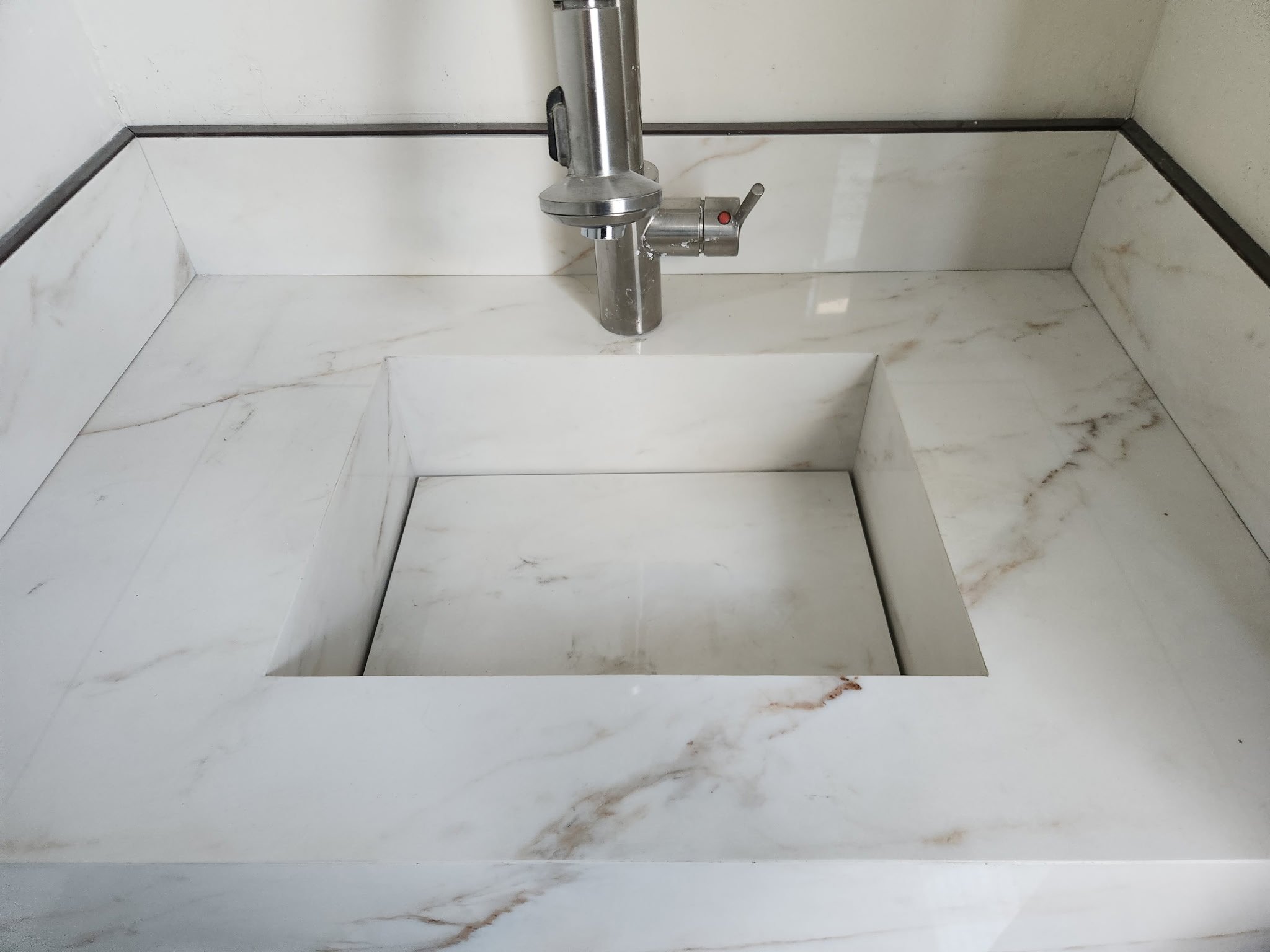 A rectangular hole in a white marble wall with brown veining, with a stainless steel pipe and valve attached near the top center of the hole.