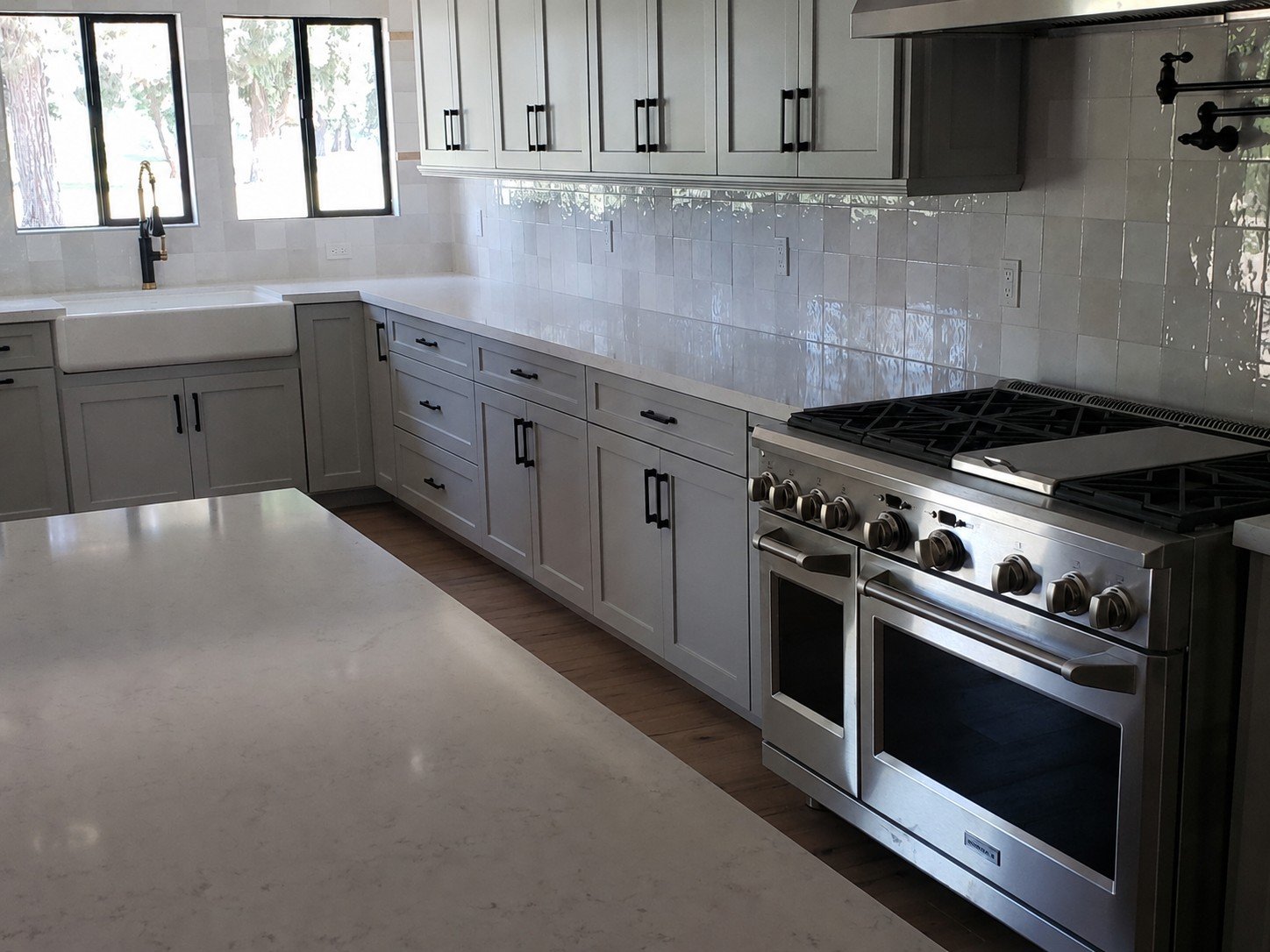 Modern kitchen with gray cabinets, white countertops, a white farmhouse sink, a black faucet, and a stainless steel stove with oven. Window above the sink shows trees outside.