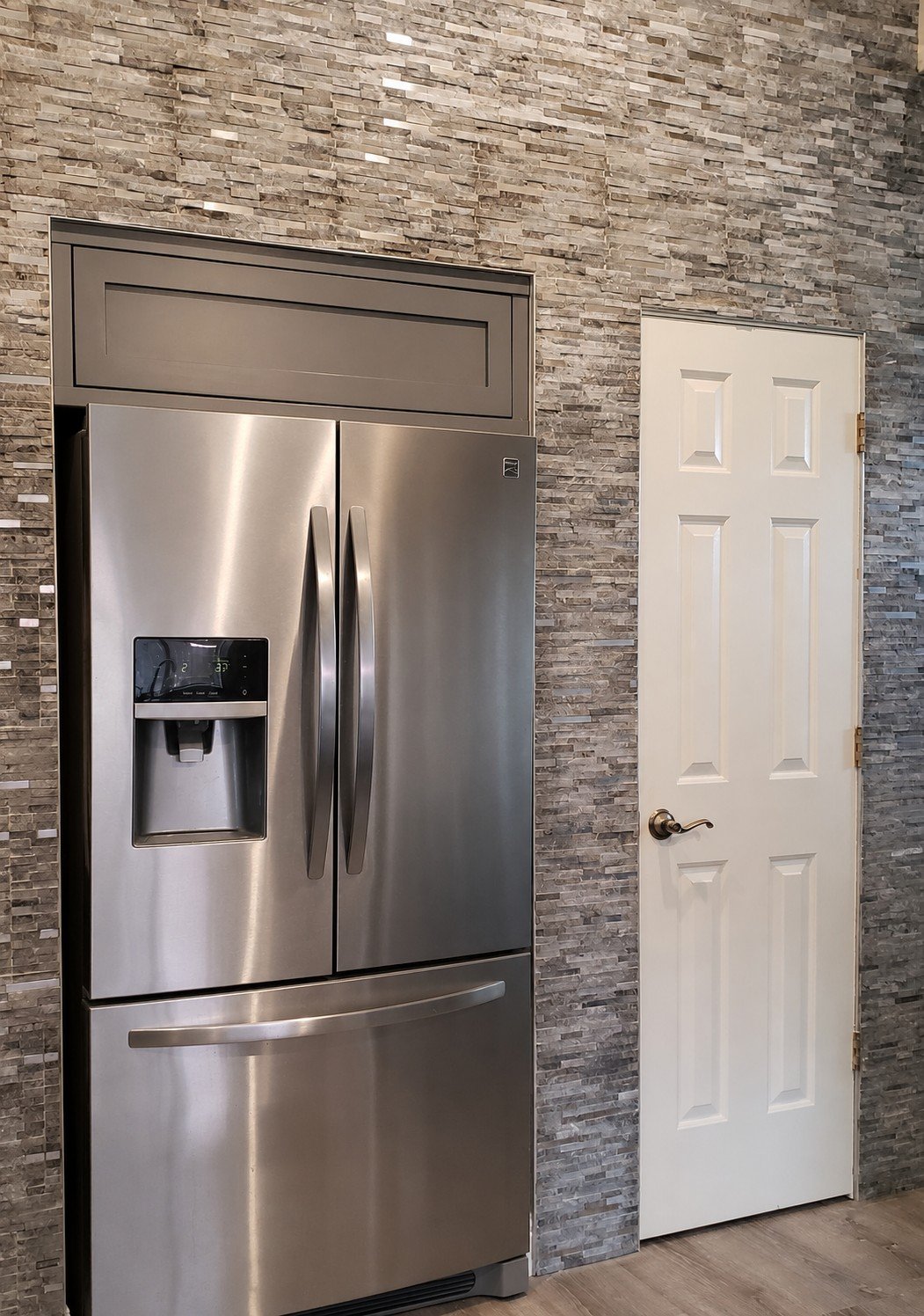 Stainless steel side-by-side refrigerator with water and ice dispenser, adjacent to a white six-panel door, against a textured gray stone wall.