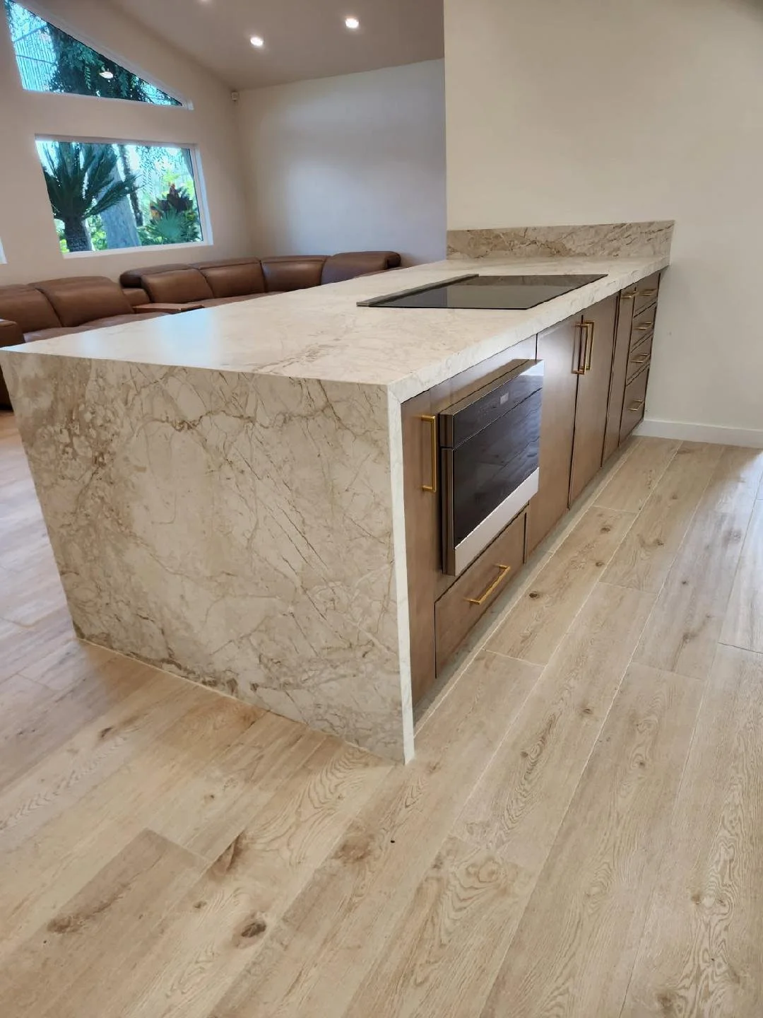 Kitchen island with beige marble countertop and wooden cabinets with gold handles, oven, and an electric cooktop, with a living room and window with plants in the background.