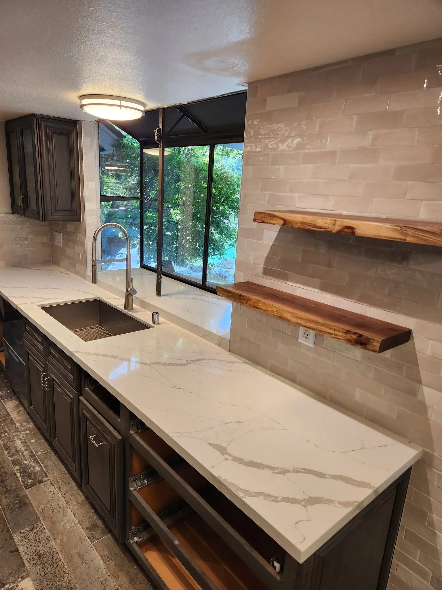Modern kitchen with white marble countertop, dark wooden cabinets, and a large window with greenery outside.