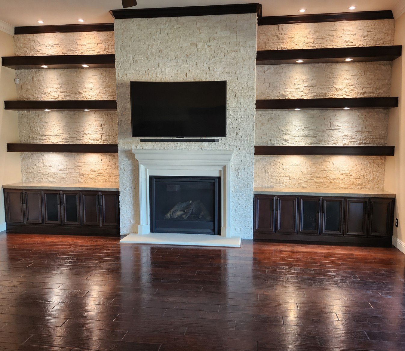 Living room with a fireplace, a mounted flat-screen TV above it, and built-in dark wood cabinets with open shelves on either side, illuminated by ceiling lights.