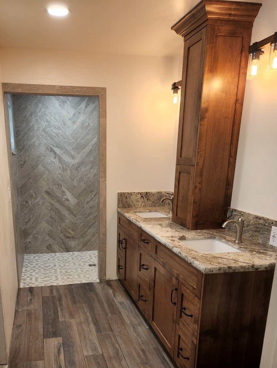 Bathroom with a wood vanity and granite countertop, two sinks, and an adjoining walk-in shower with gray tiled walls and a mosaic tile floor.