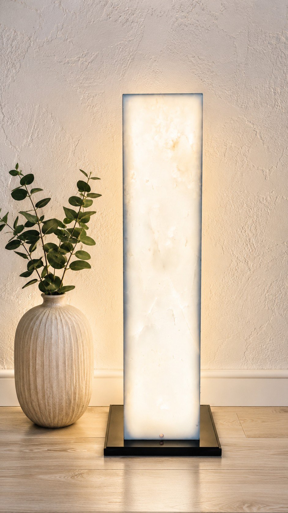 A tall, rectangular, backlit lamp with a cream-colored marble-like surface, placed on a wooden floor against a beige textured wall. To the left, a beige ceramic vase with vertical ridges contains a leafy green plant.