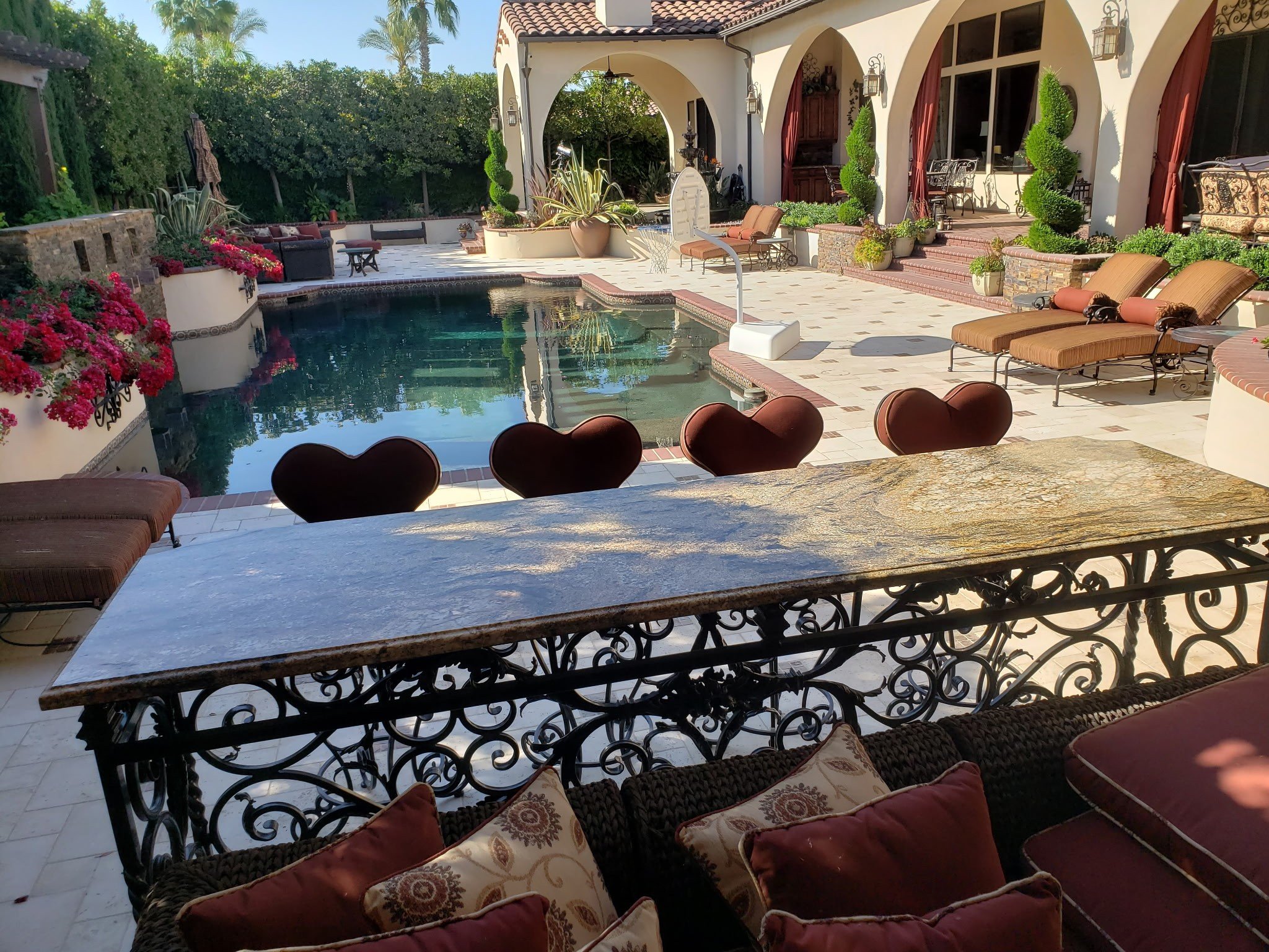 A backyard patio with a swimming pool, surrounded by potted plants, outdoor furniture, and a covered veranda with arches and red curtains. The area features lounge chairs and decorative landscaping, including flowering plants and greenery.