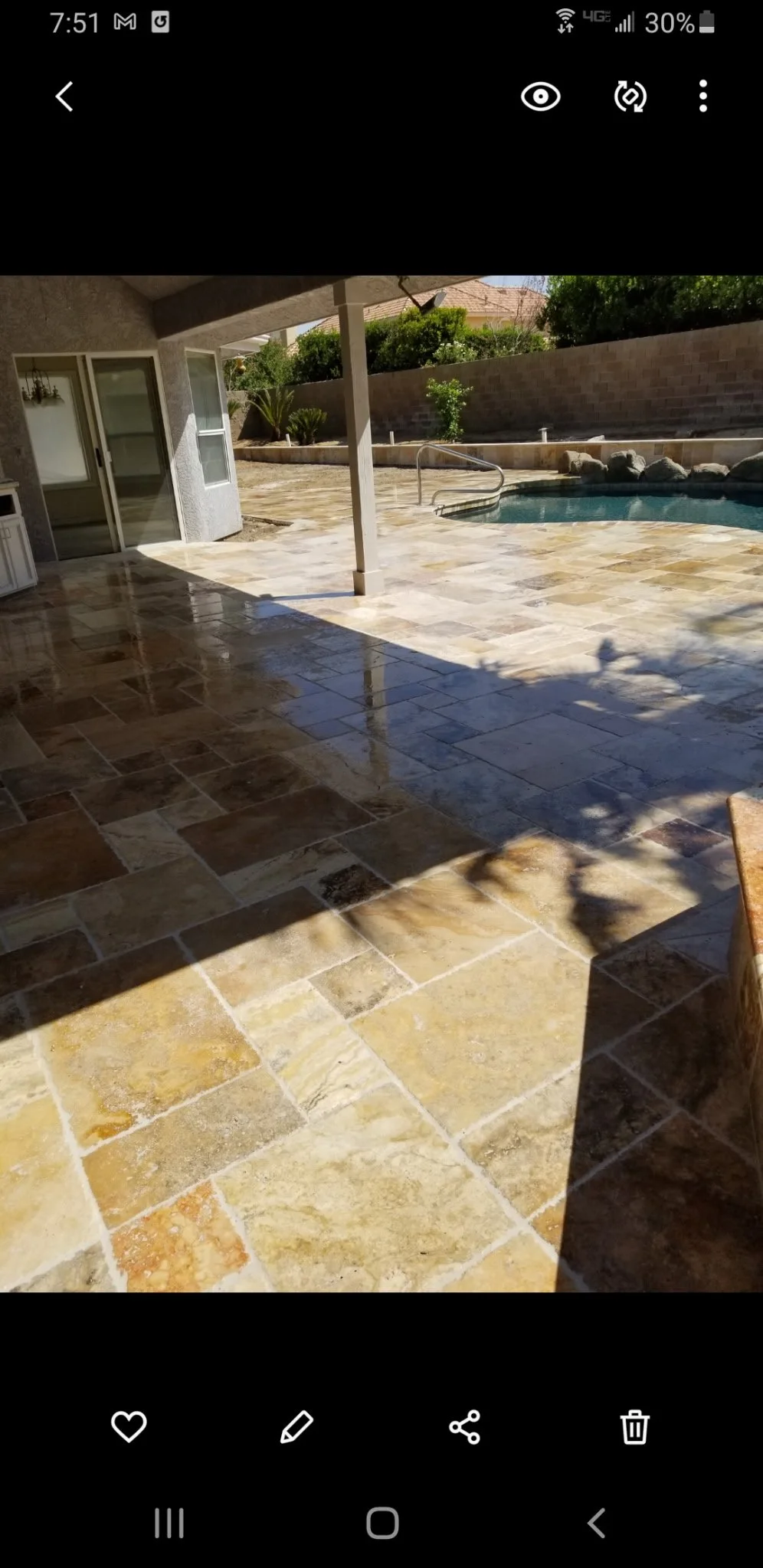 Backyard patio with tan stone tiles and a swimming pool, shaded area, and greenery in the background.