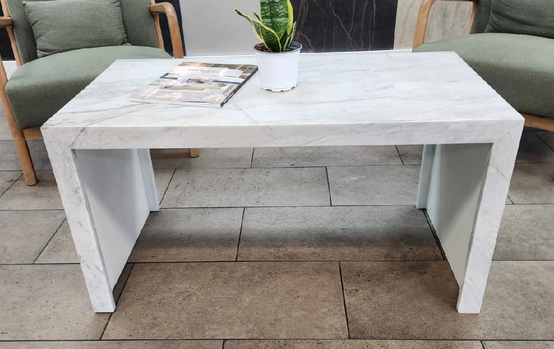 A white marble coffee table with a potted snake plant and magazines on top, surrounded by green cushioned chairs on a tiled floor.