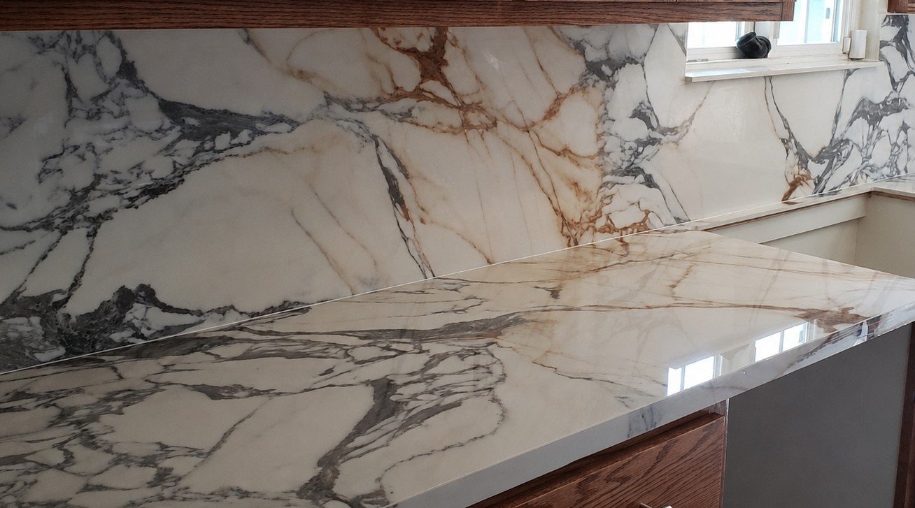 A marble kitchen countertop and backsplash in shades of white, gray, and brown, with a window above.