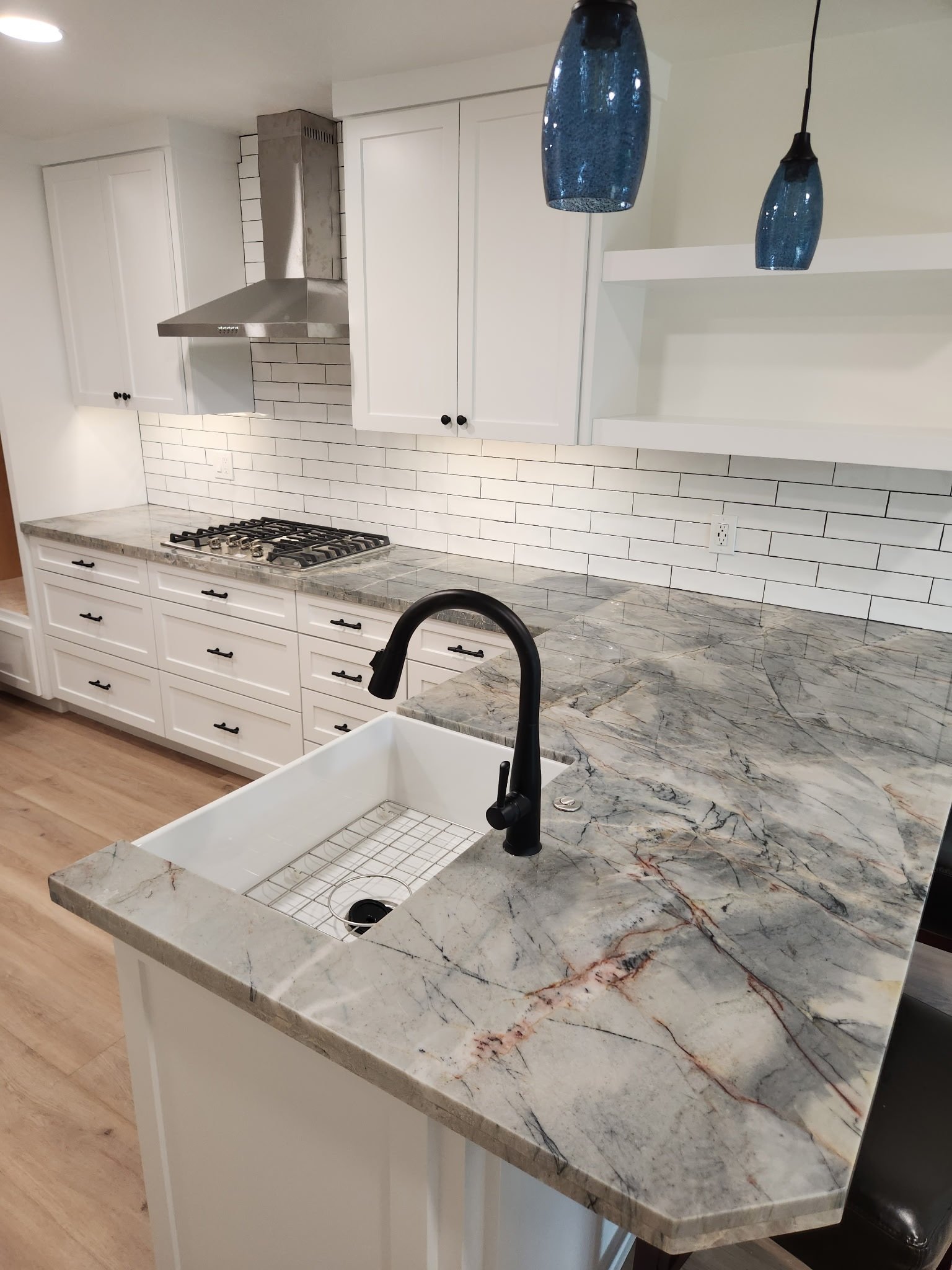 A kitchen with white cabinets, a marble countertop with gray, white, and reddish veins, a white farmhouse sink with a black faucet, a white tile backsplash with black grout, a stainless steel range hood, and two blue pendant lights hanging from the ceiling.