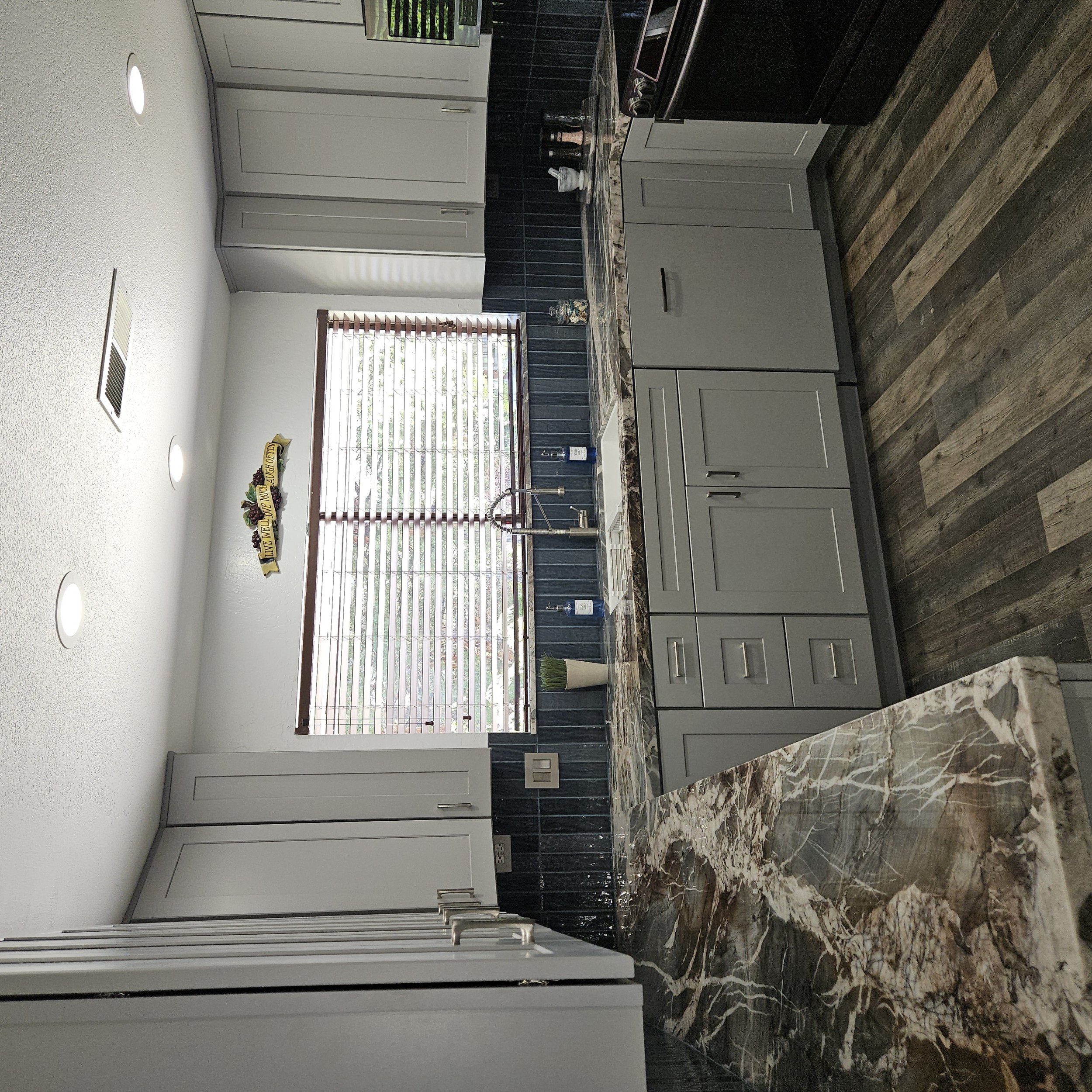 Modern kitchen with gray cabinets, marble countertop, black backsplash, and wooden flooring, illuminated by ceiling lights and natural light through blinds.