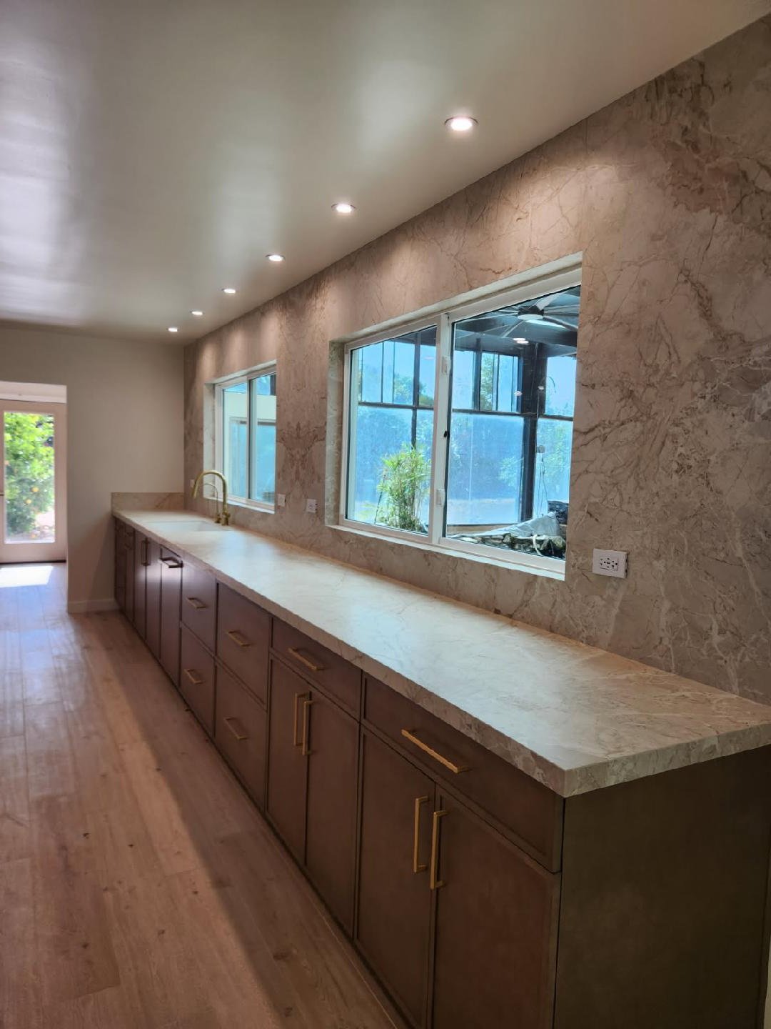 Modern kitchen with a long marble countertop, wooden cabinets with gold handles, and large windows overlooking an outdoor area with plants.