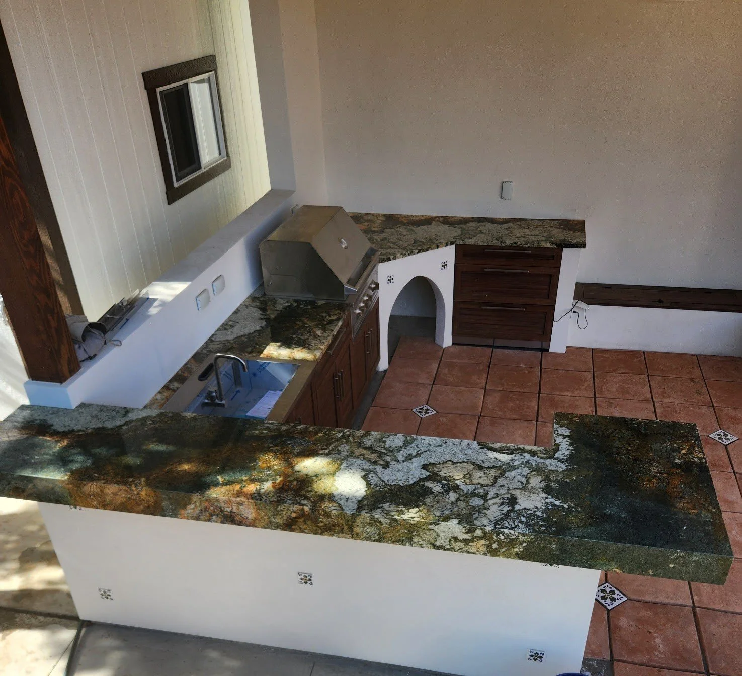 Kitchen with granite countertops, a stainless steel stove, a sink with a faucet, wooden cabinets, a small arched space, and terracotta tile floors.