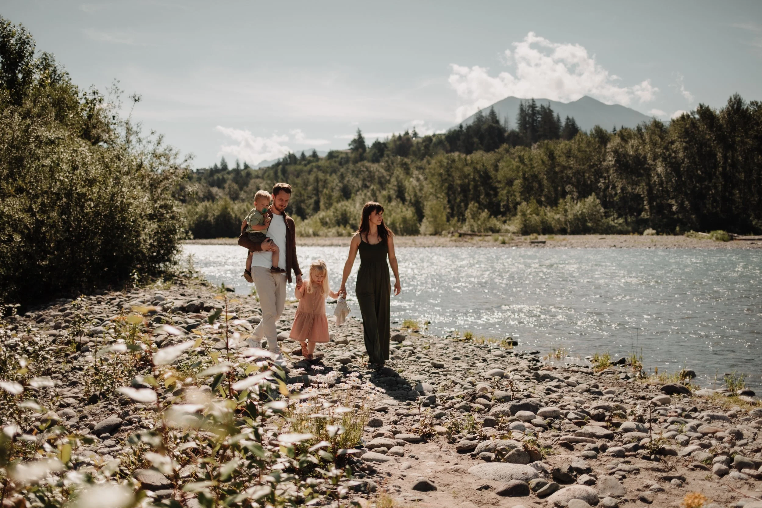 A family walking along a rocky riverbank with mountains and trees in the background on a sunny day.