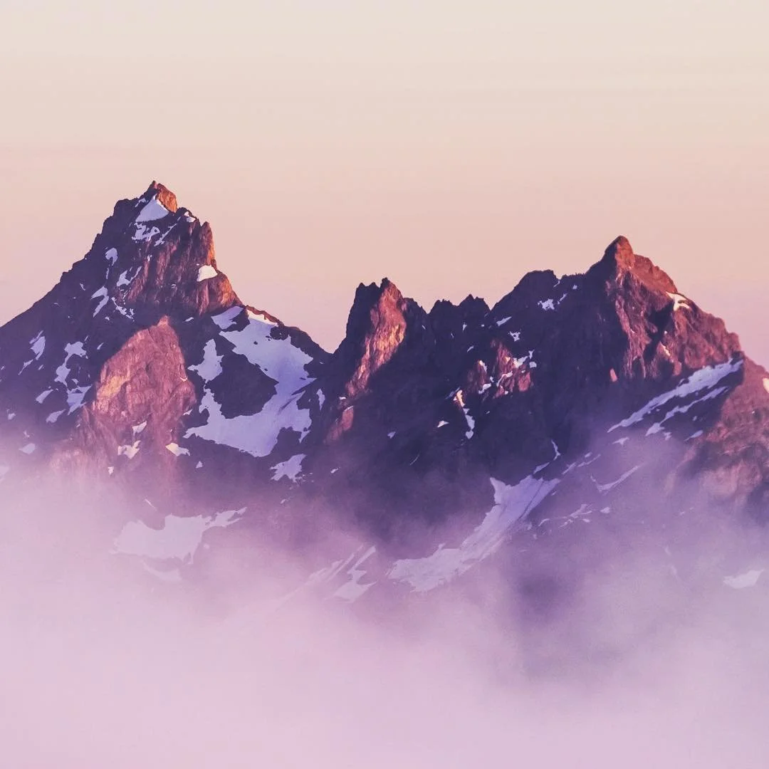Snow-capped mountain peaks emerging above fog with a pinkish sky.