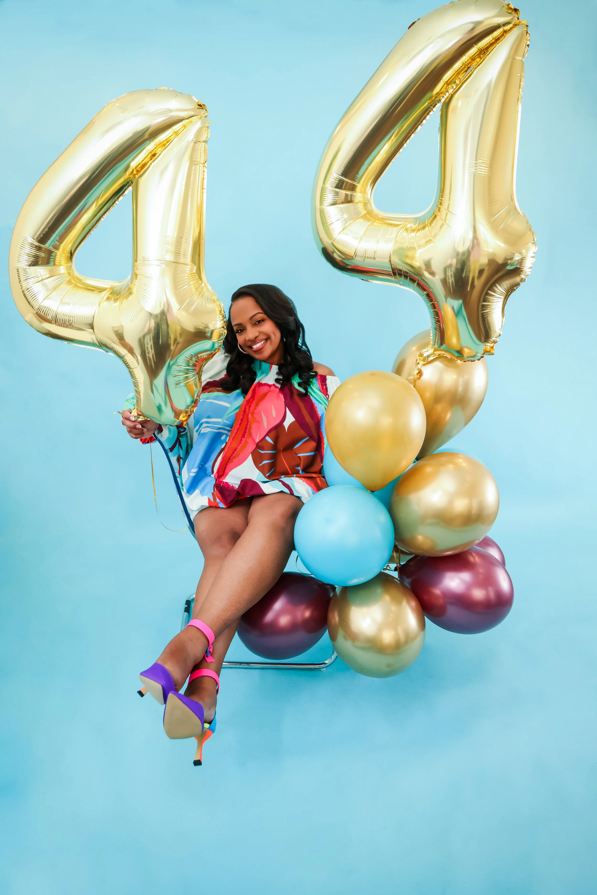 A woman sitting on a chair in front of a blue background, holding two large gold balloons shaped as the number four, and surrounded by numerous colorful balloons, wearing a colorful dress and high heels.