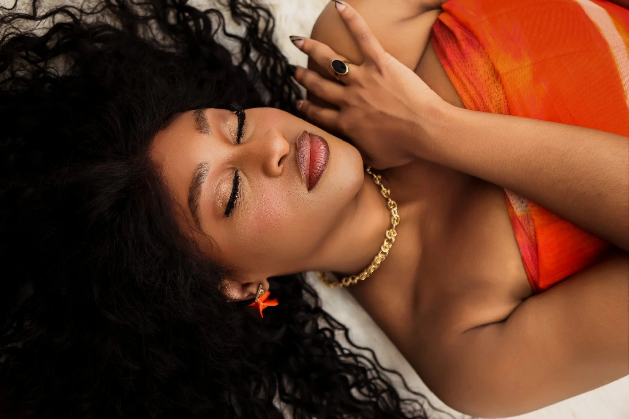 A woman lying down with closed eyes, dark curly hair, wearing orange clothing and gold jewelry, with one hand resting on her chest.
