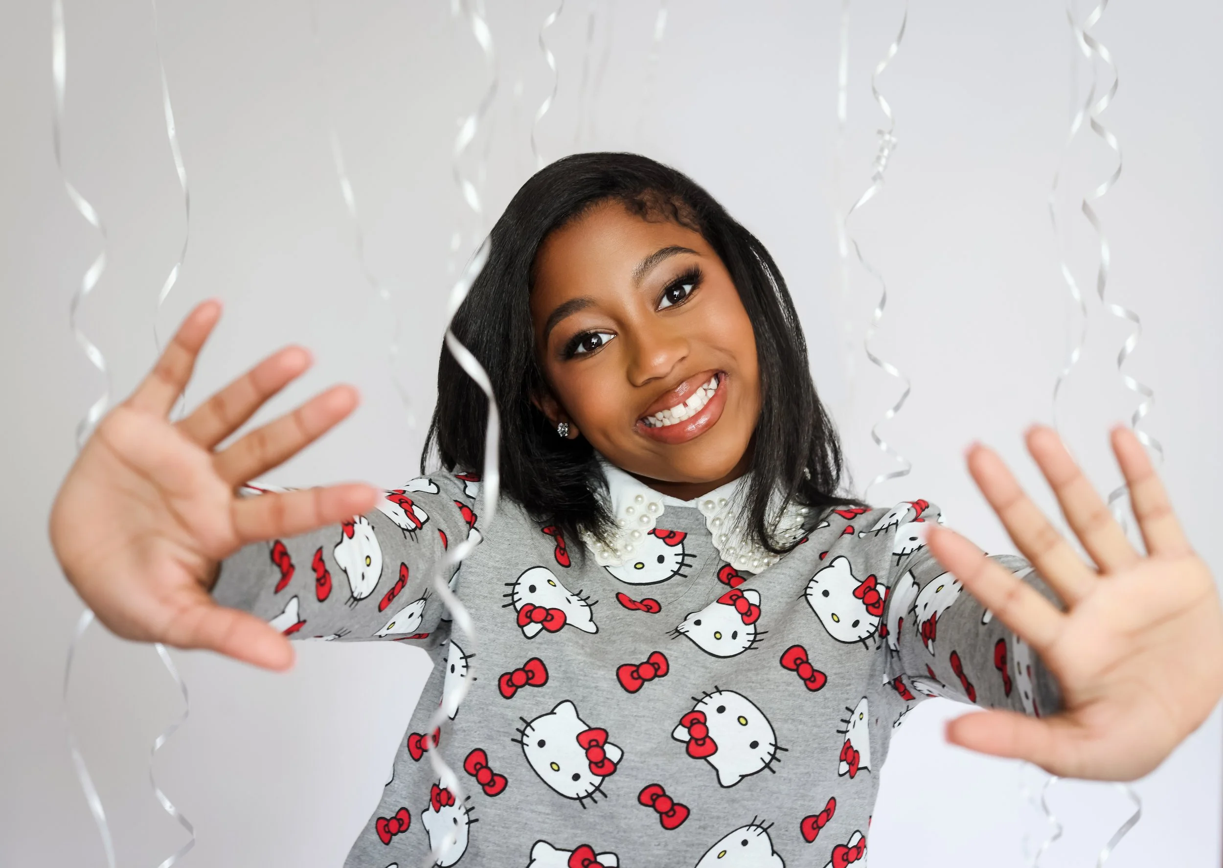 A young woman smiling and reaching towards the camera, with metallic curly ribbons hanging in the foreground. She is wearing a grey Hello Kitty sweatshirt with a pearl collar.