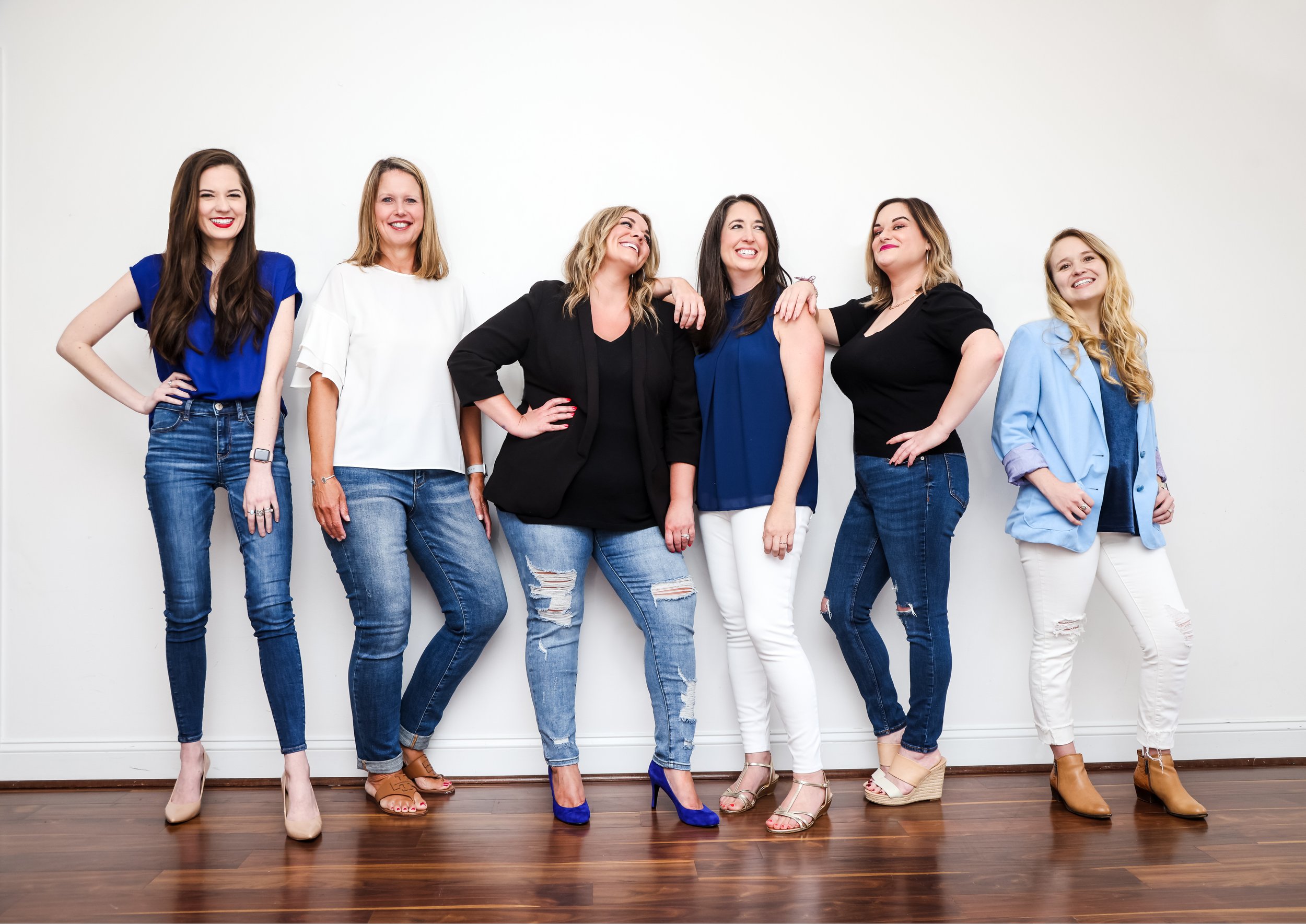 Six women standing in a line against a plain white wall, smiling and interacting with each other, dressed in casual and semi-formal outfits.