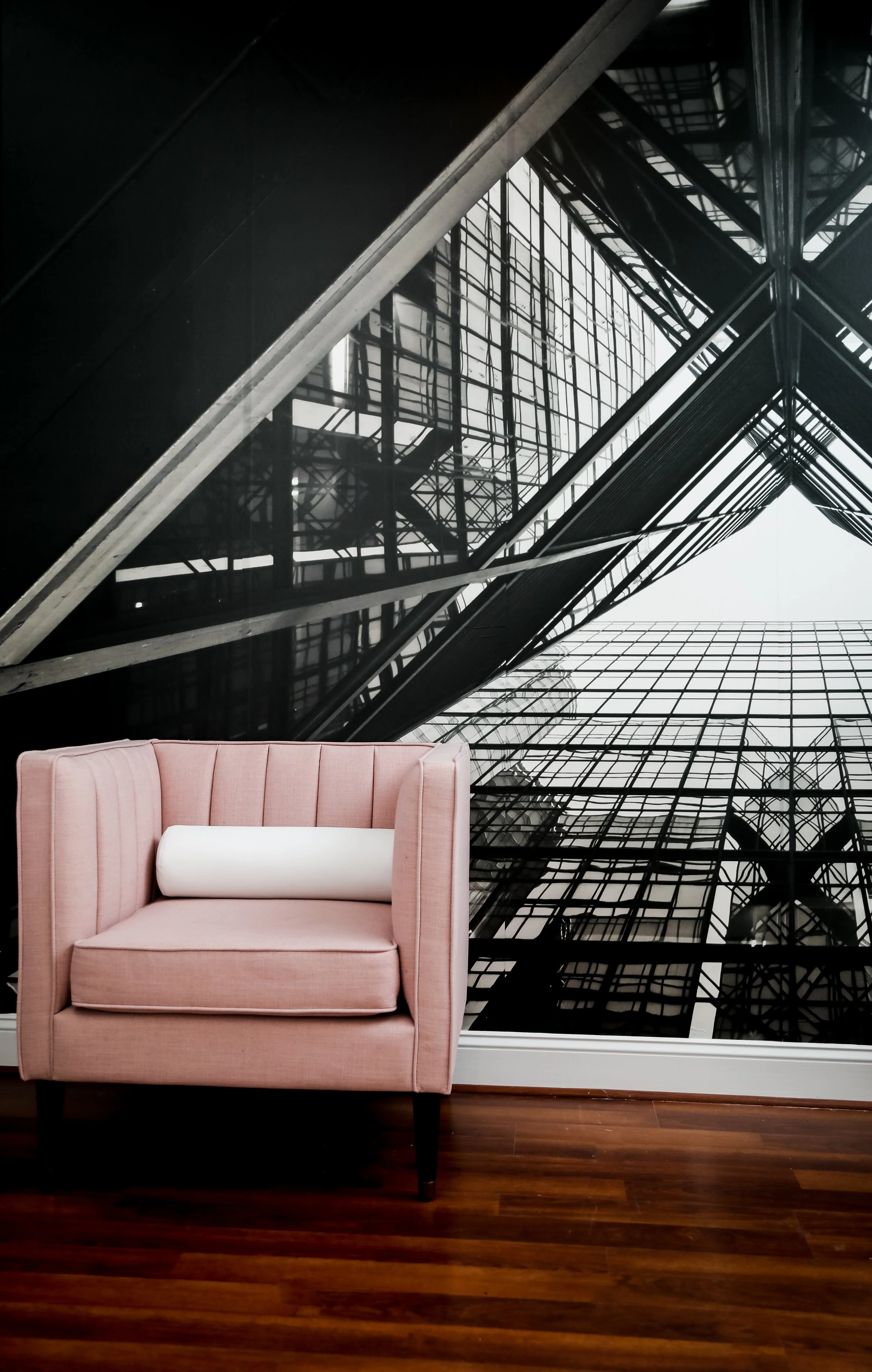 A pink upholstered armchair with a white cylindrical pillow on a hardwood floor, placed in front of a large black and white photograph of a modern building with glass and steel architecture.