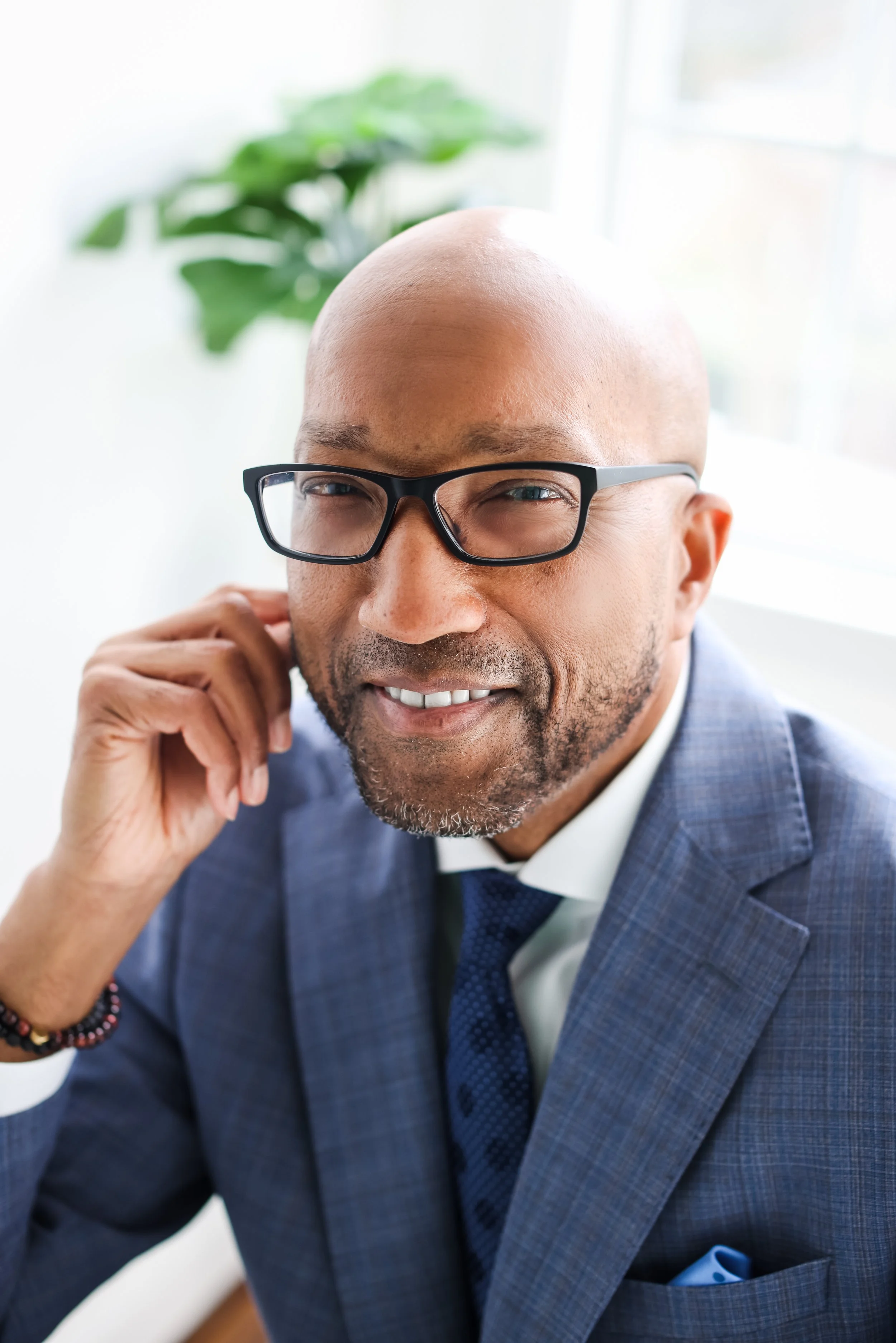 Close-up of a smiling African American man with glasses, dressed in a blue suit and white shirt, holding his hand near his face, with a blurred green plant in the background.