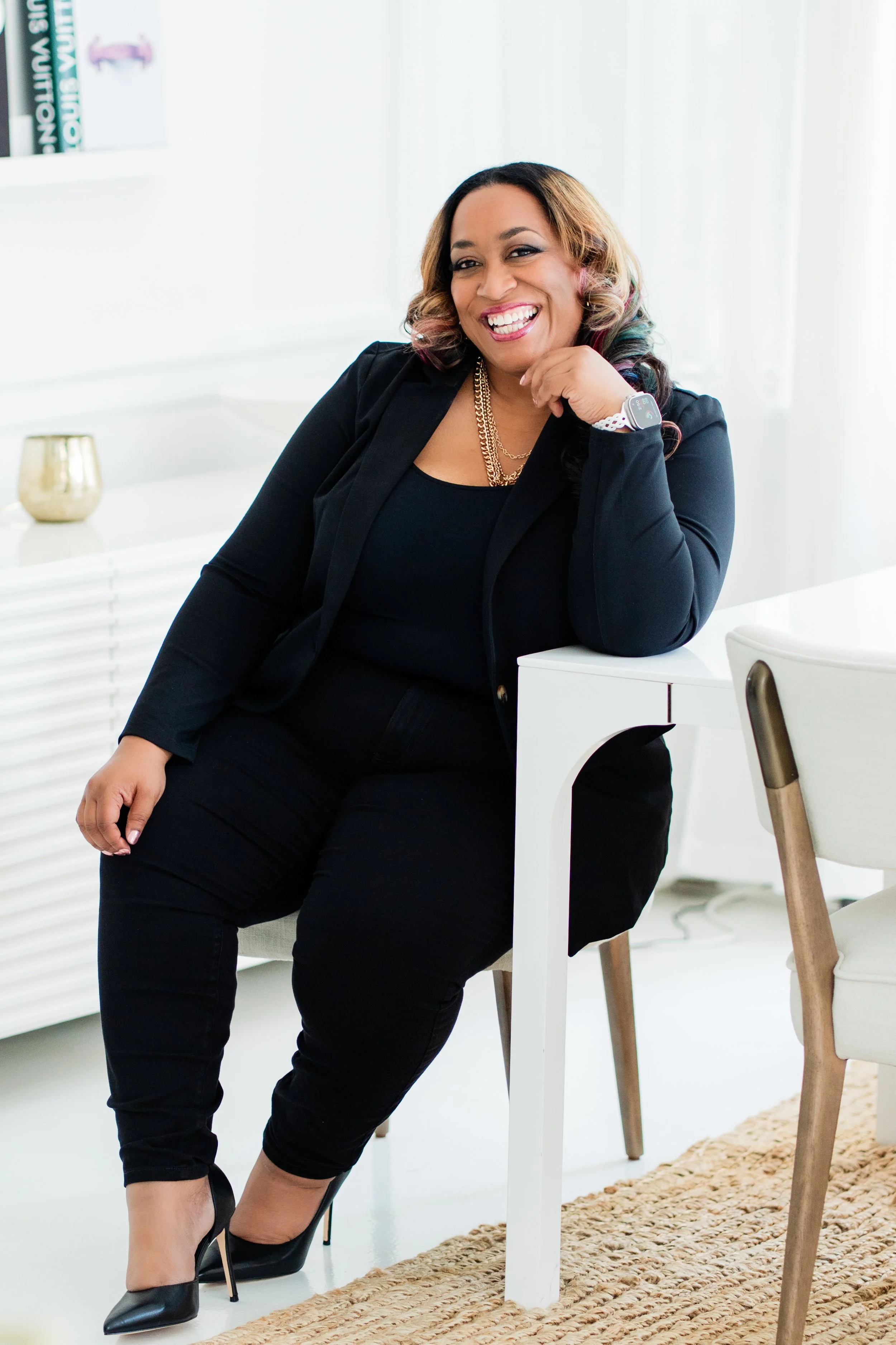 A smiling woman in a black blazer and pants sitting at a white table, wearing black high heels and a smartwatch, in a bright, modern room with white walls, a bookshelf, and a rug.