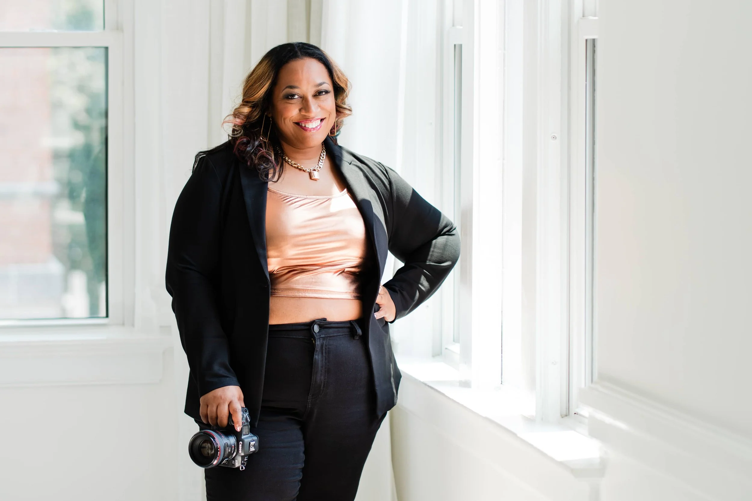 A woman standing indoors near a window, holding a professional camera, smiling, wearing a black blazer and pants, with a pink top and a necklace.