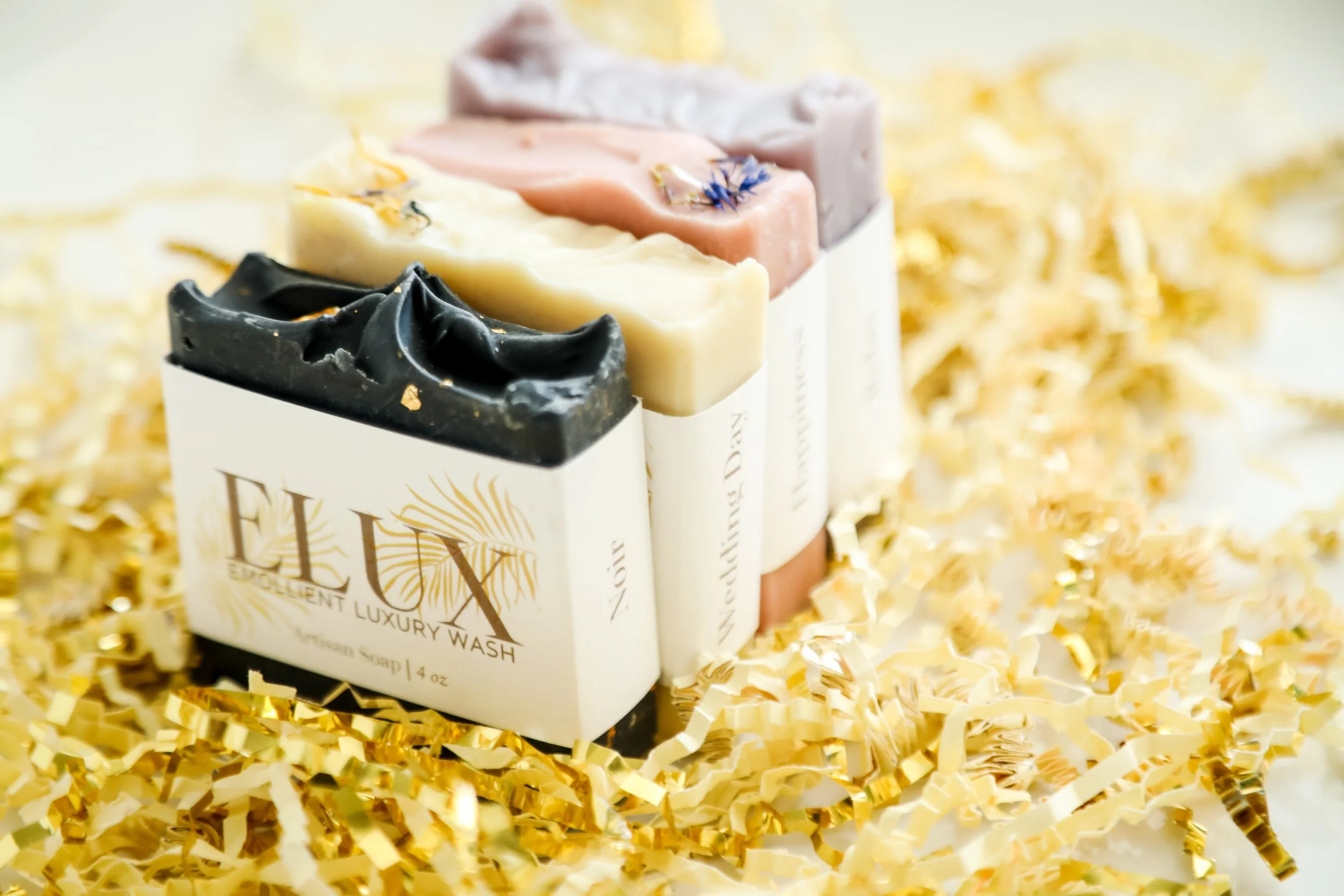 Assorted handmade soap bars in black, white, pink, and purple, displayed on a white box with golden foil confetti in the background.