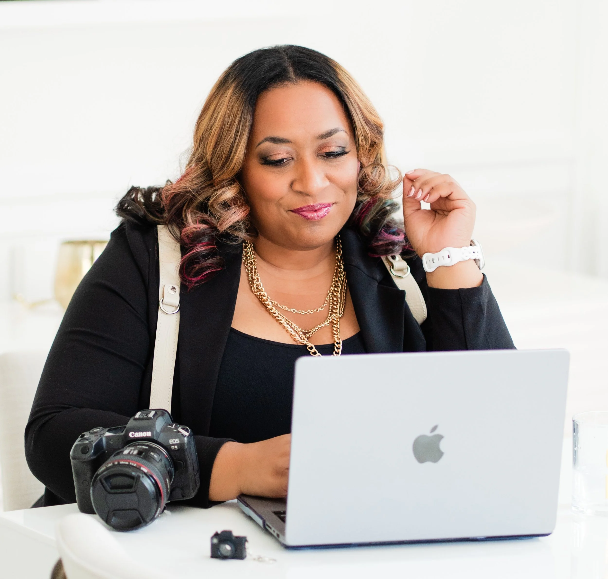 A woman with shoulder-length hair sitting at a white table, looking at her laptop with a slight smile. There is a Canon DSLR camera with a telephoto lens on the table, and a small camera accessory in front of her. The background is blurred and appears to be a bright room.