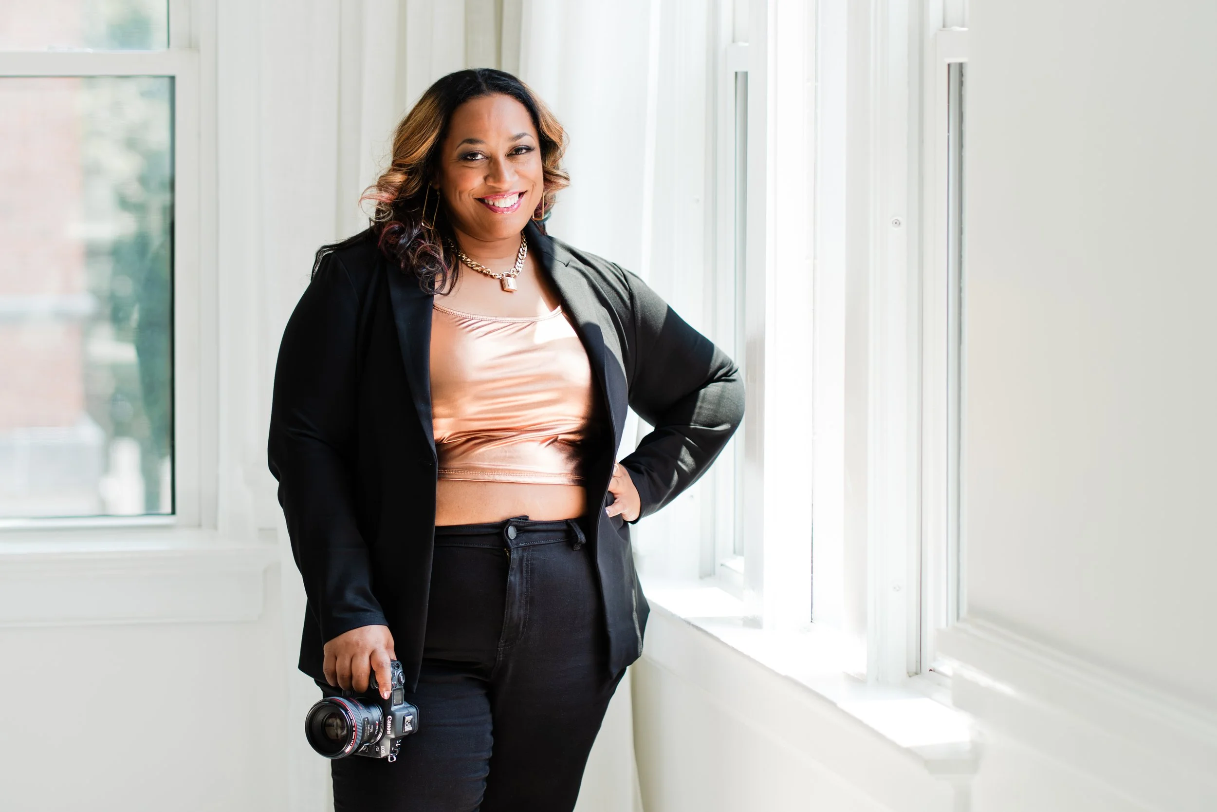 A confident woman standing by a large window, holding a professional camera in her right hand, dressed in a black blazer and pants, smiling in a well-lit room.