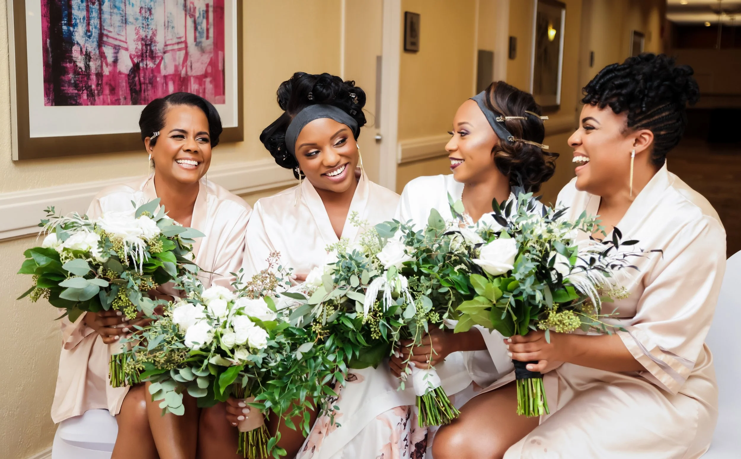 Four women dressed in satin robes holding bouquets of white flowers and greenery, smiling and enjoying a friendly moment in a hallway with abstract artwork on the wall.