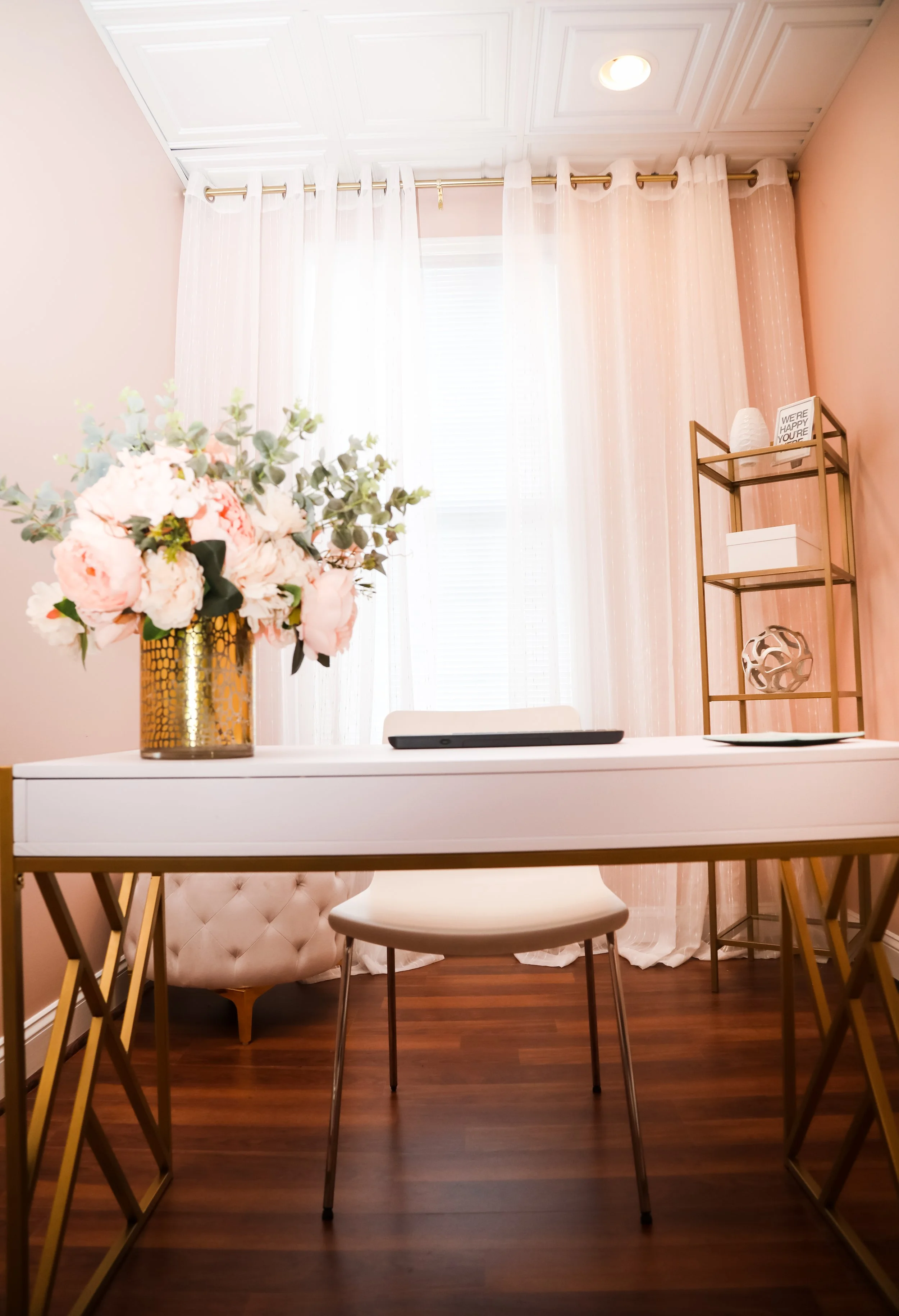 A bright room with pink and white walls, sheer curtains letting in natural light, a modern decor shelf with decor items and a framed quote, and a table with a floral arrangement in a gold vase.