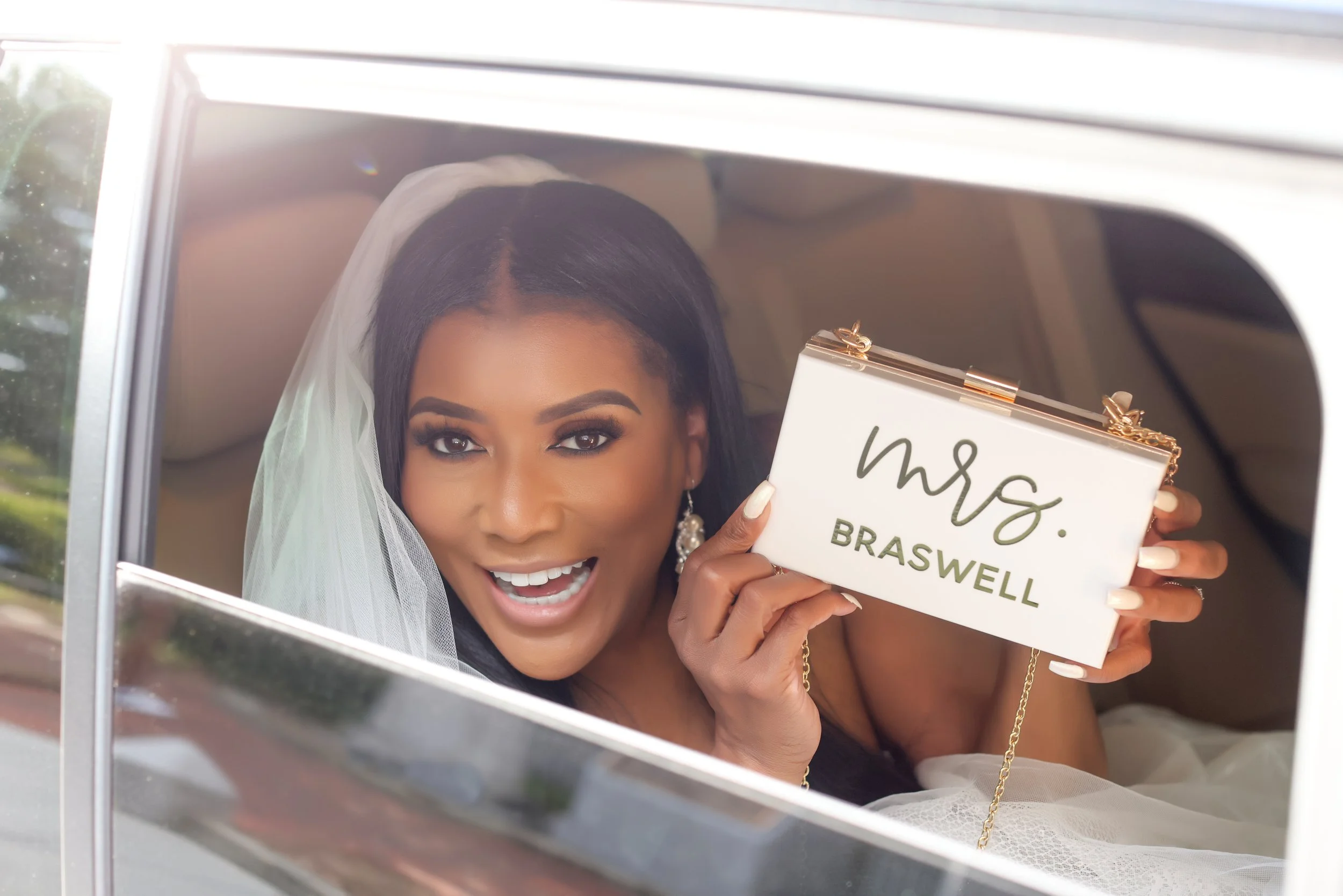 A smiling woman with dark hair, wearing makeup and earrings, is holding a sign that reads 'Mrs. Braswell' inside a vehicle. She is wearing a white veil, indicating she is a bride, and looking through a window at the camera.