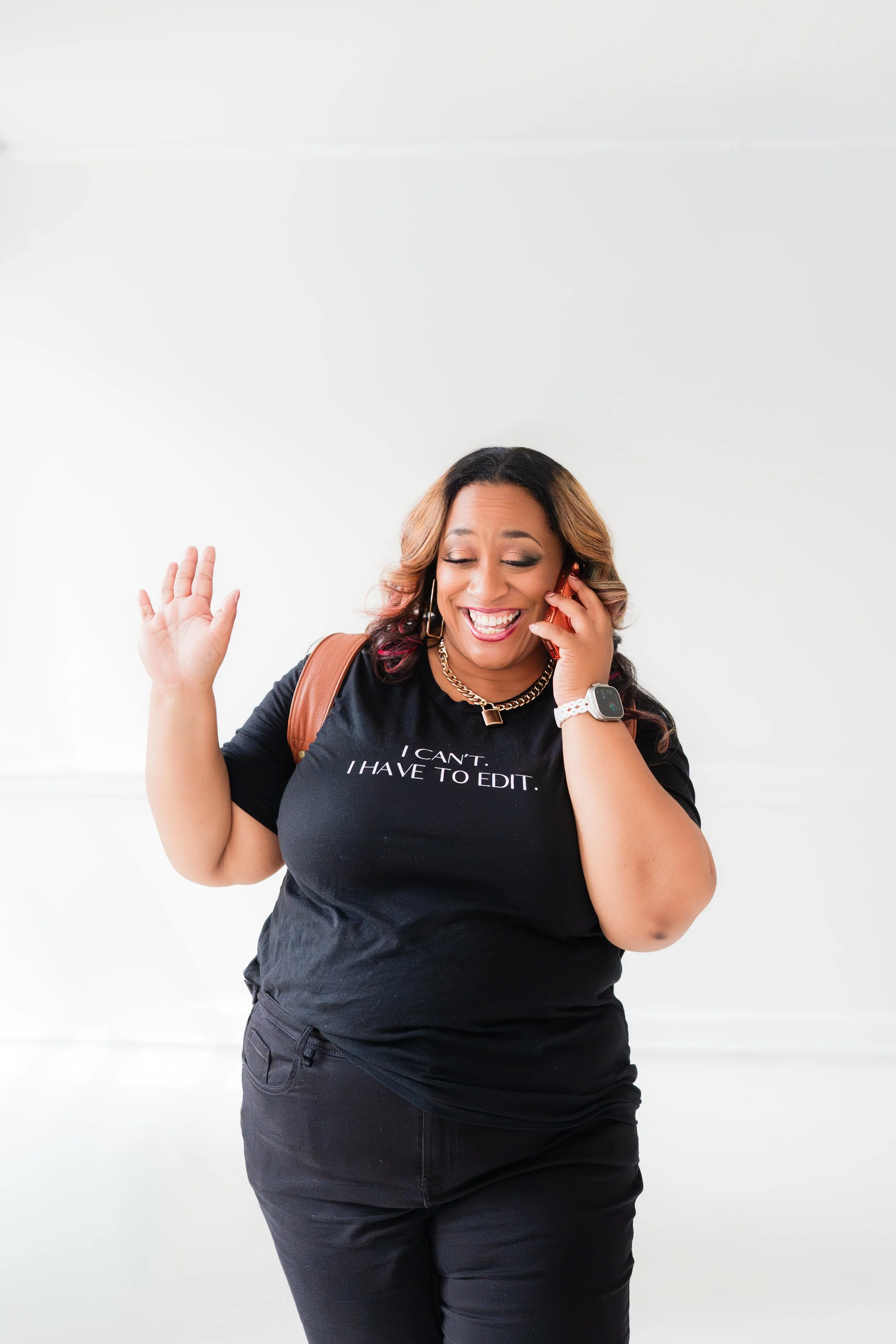 A woman smiling and talking on a cell phone, wearing a black t-shirt with the text 'I CAN'T. I HAVE TO EDIT.' and a backpack, against a plain white background.