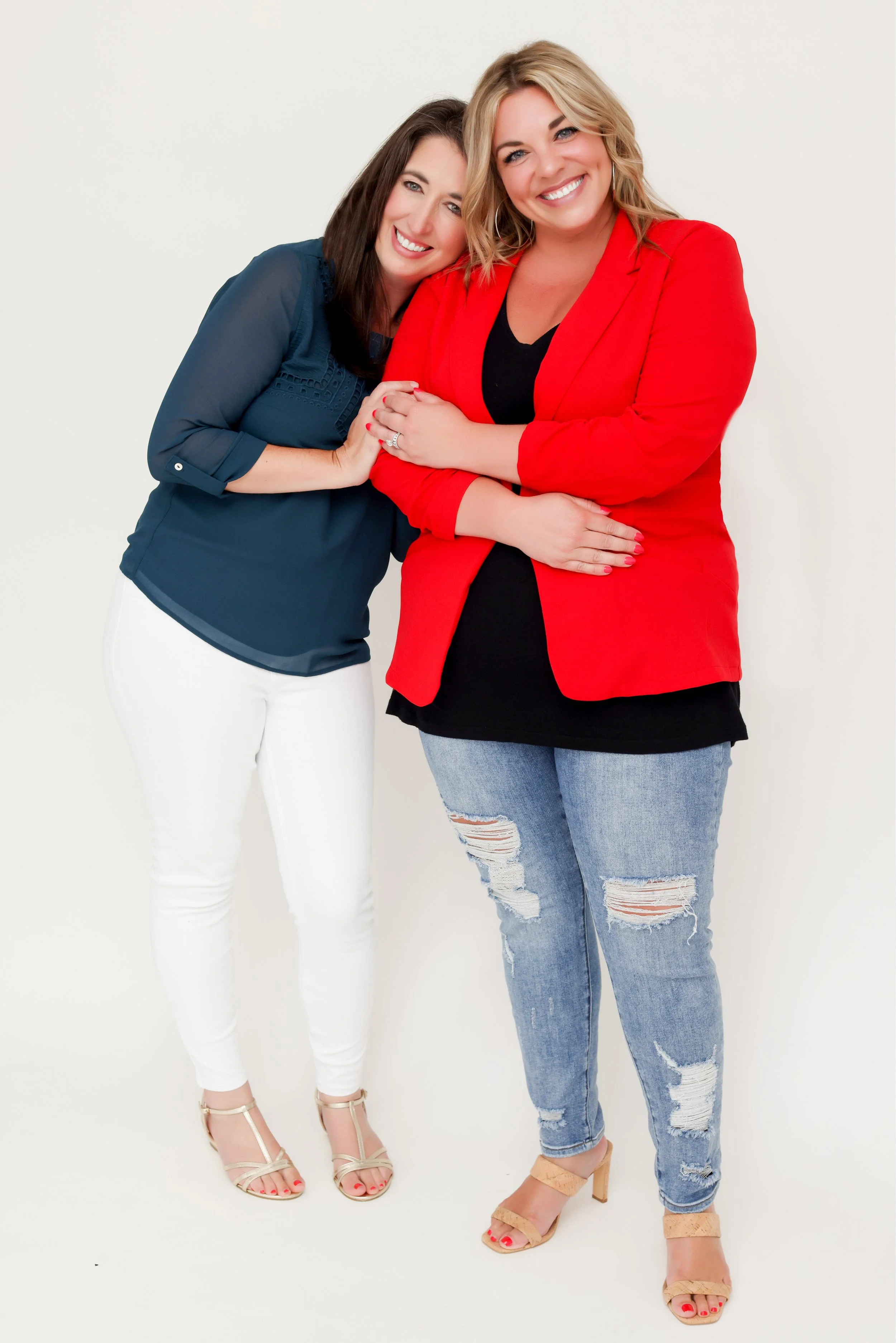 Two women smiling and posing together. The woman on the left has dark brown hair, is wearing a navy blue blouse and white pants with beige heels. The woman on the right has blonde hair, is wearing a red blazer, black top, distressed blue jeans, and beige high heels.