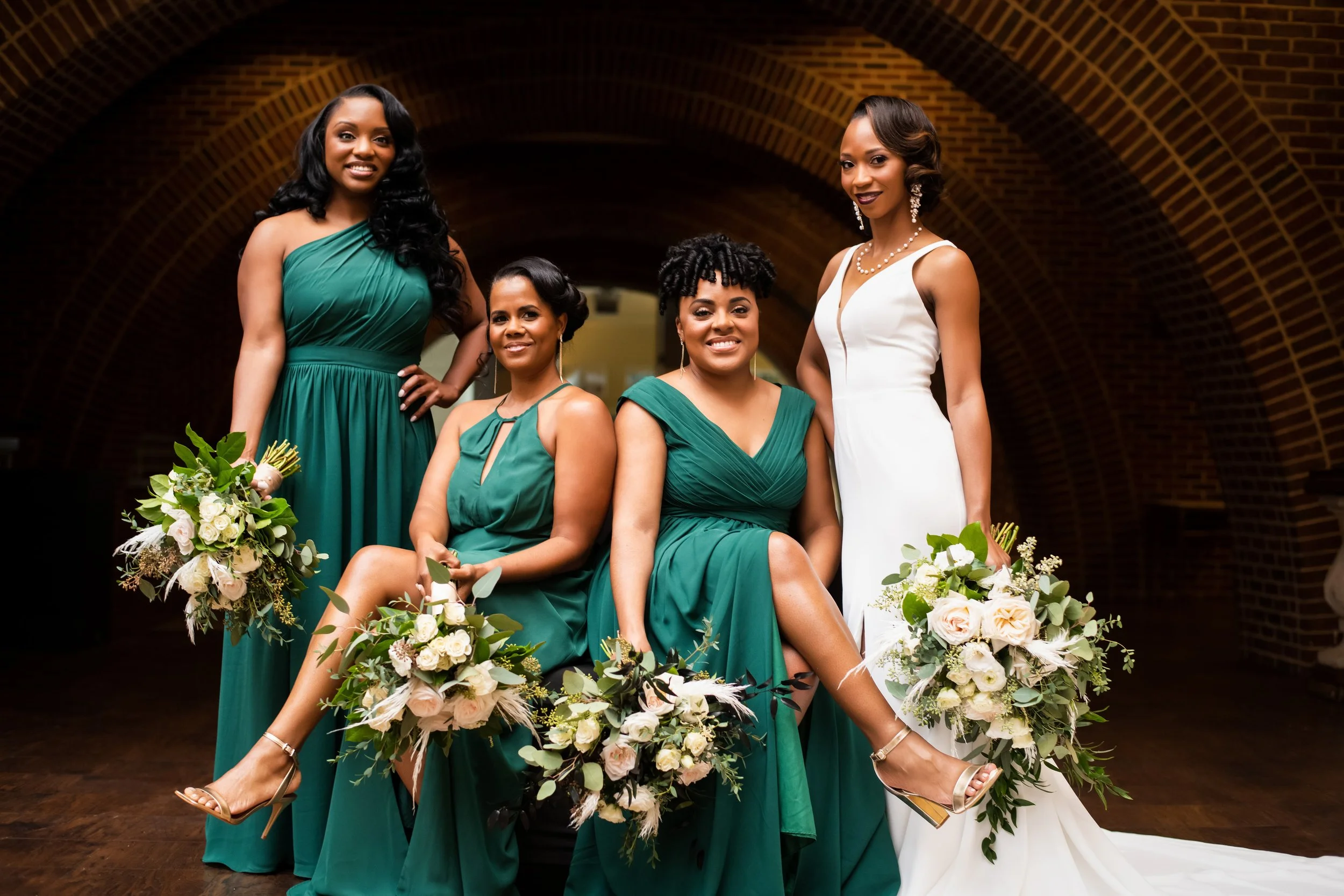 Group of five women in elegant dresses, with two women sitting and three standing, holding bouquets of flowers, posing in front of a brick archway for a formal or wedding photo.