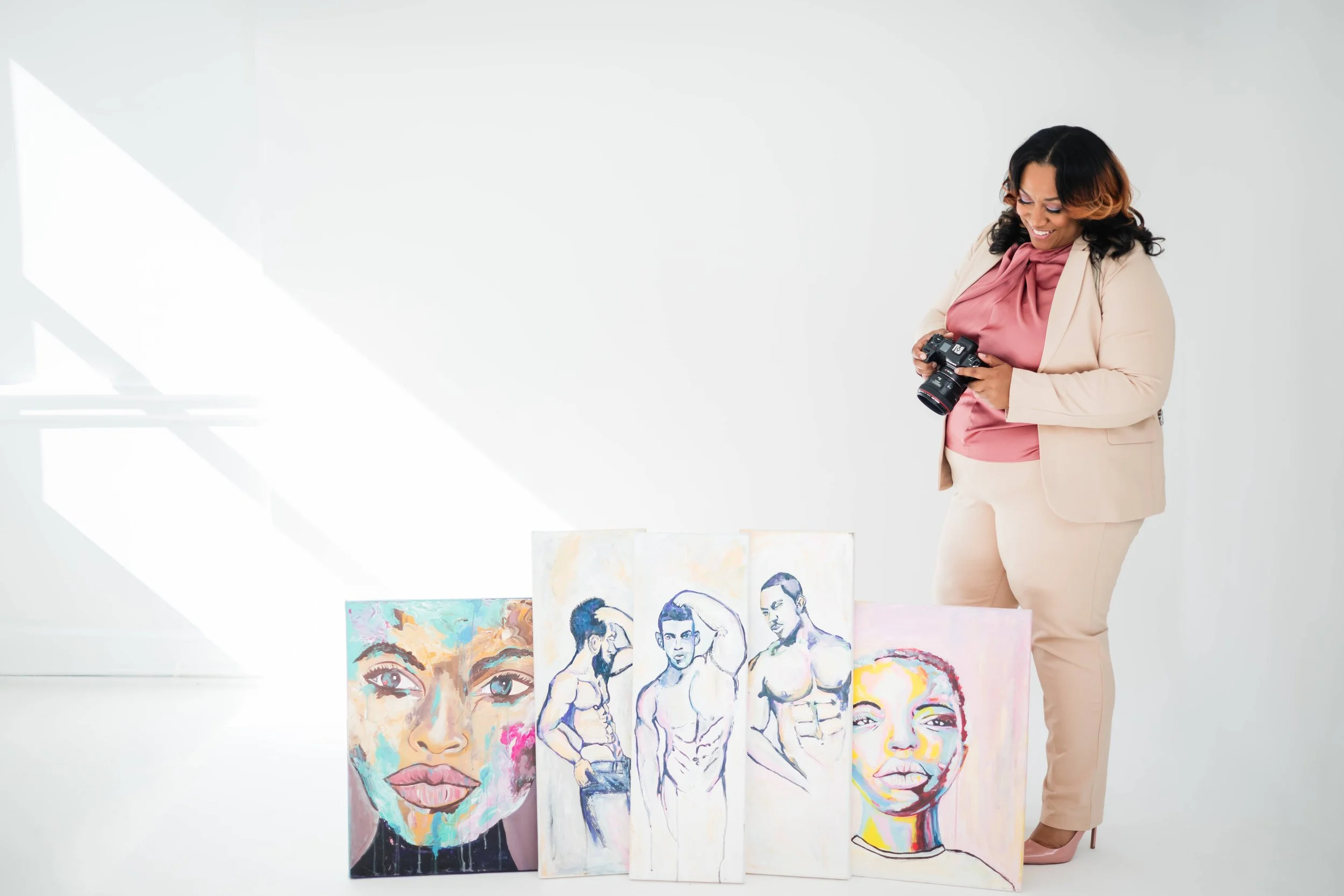 A woman in a beige suit and pink blouse holding a camera, standing next to colorful paintings of women's faces and figures in an art gallery.