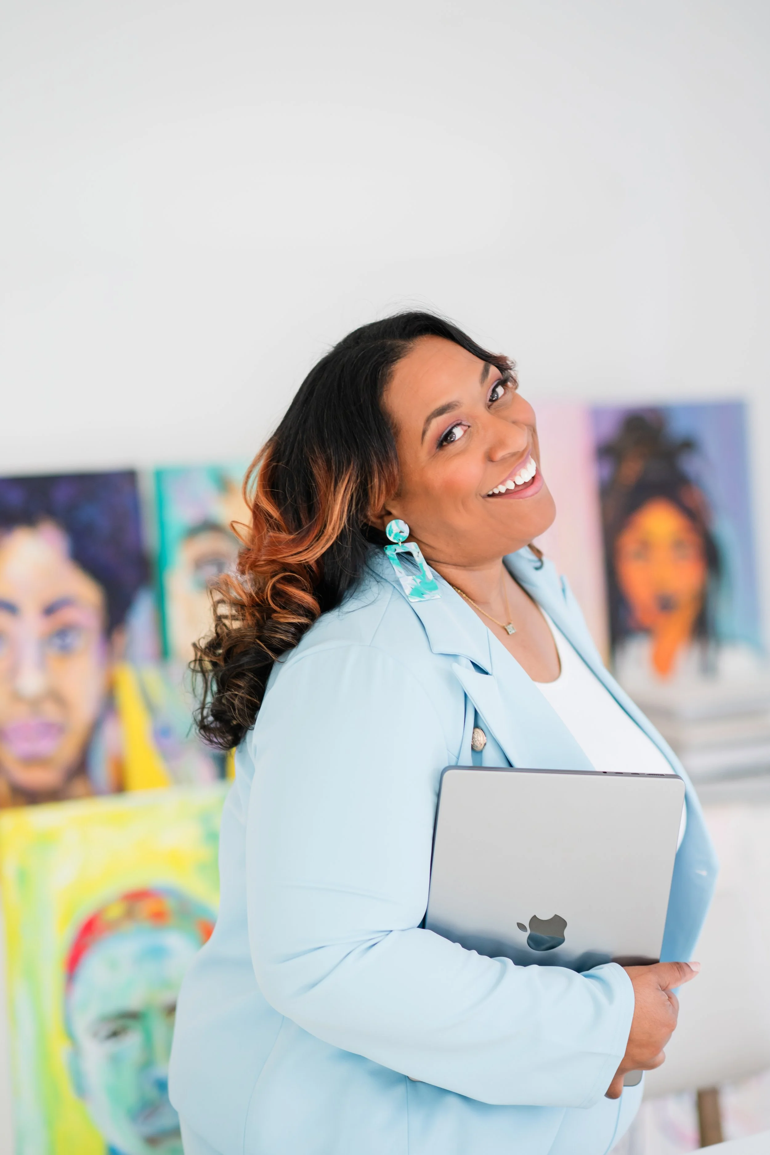 A woman with dark, wavy hair and earrings stands in an art gallery, smiling while holding an Apple MacBook Pro. Behind her are colorful paintings of faces.