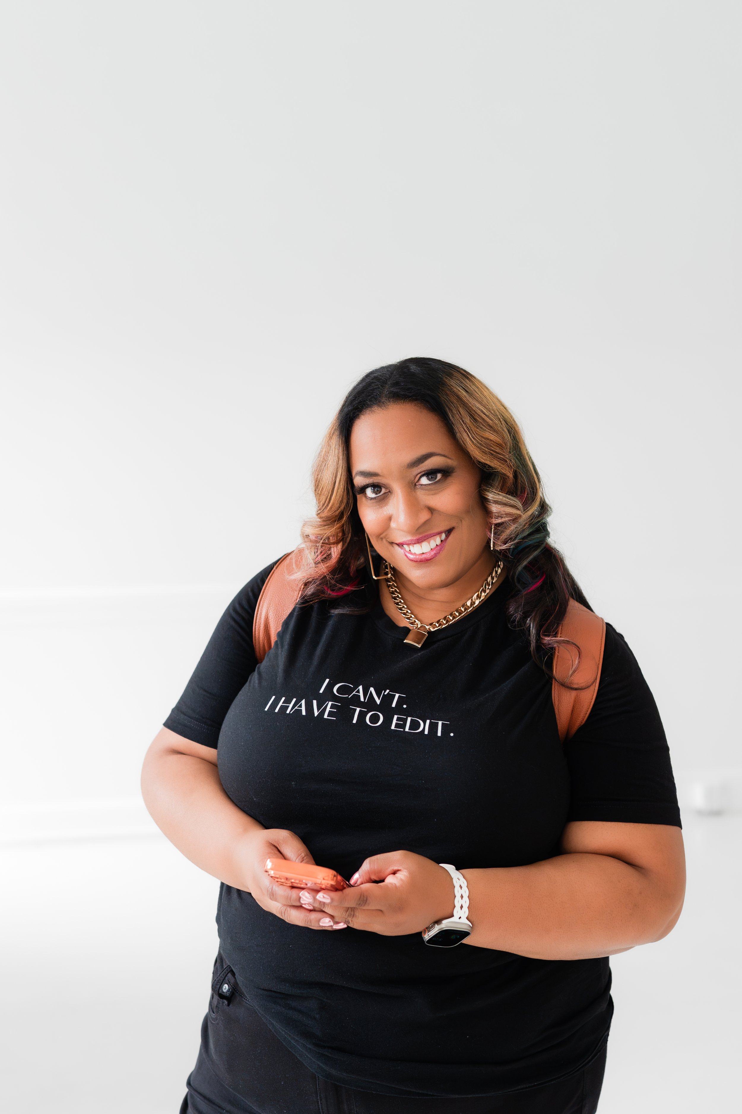 A smiling woman with wavy, multicolored hair, wearing a black T-shirt with white text, a gold chain necklace, and a white bracelet, holding a phone against a plain white background.