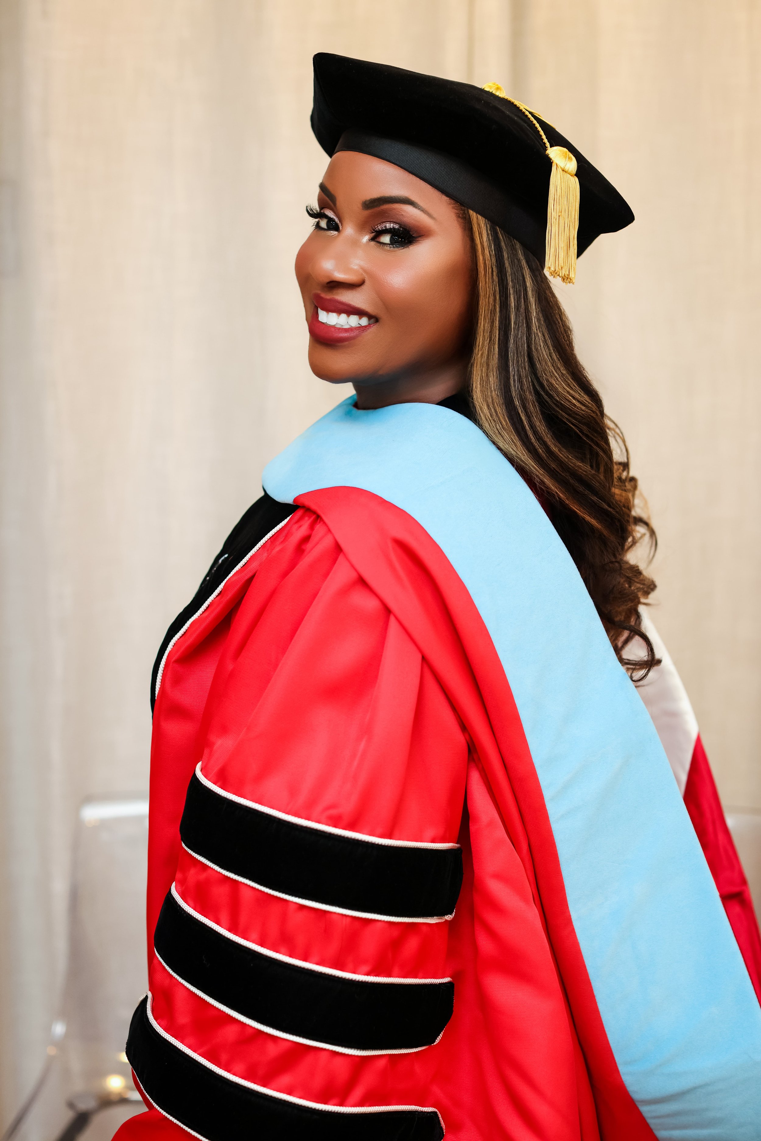 A woman dressed in a graduation cap and gown, smiling, with a light-colored curtain background.