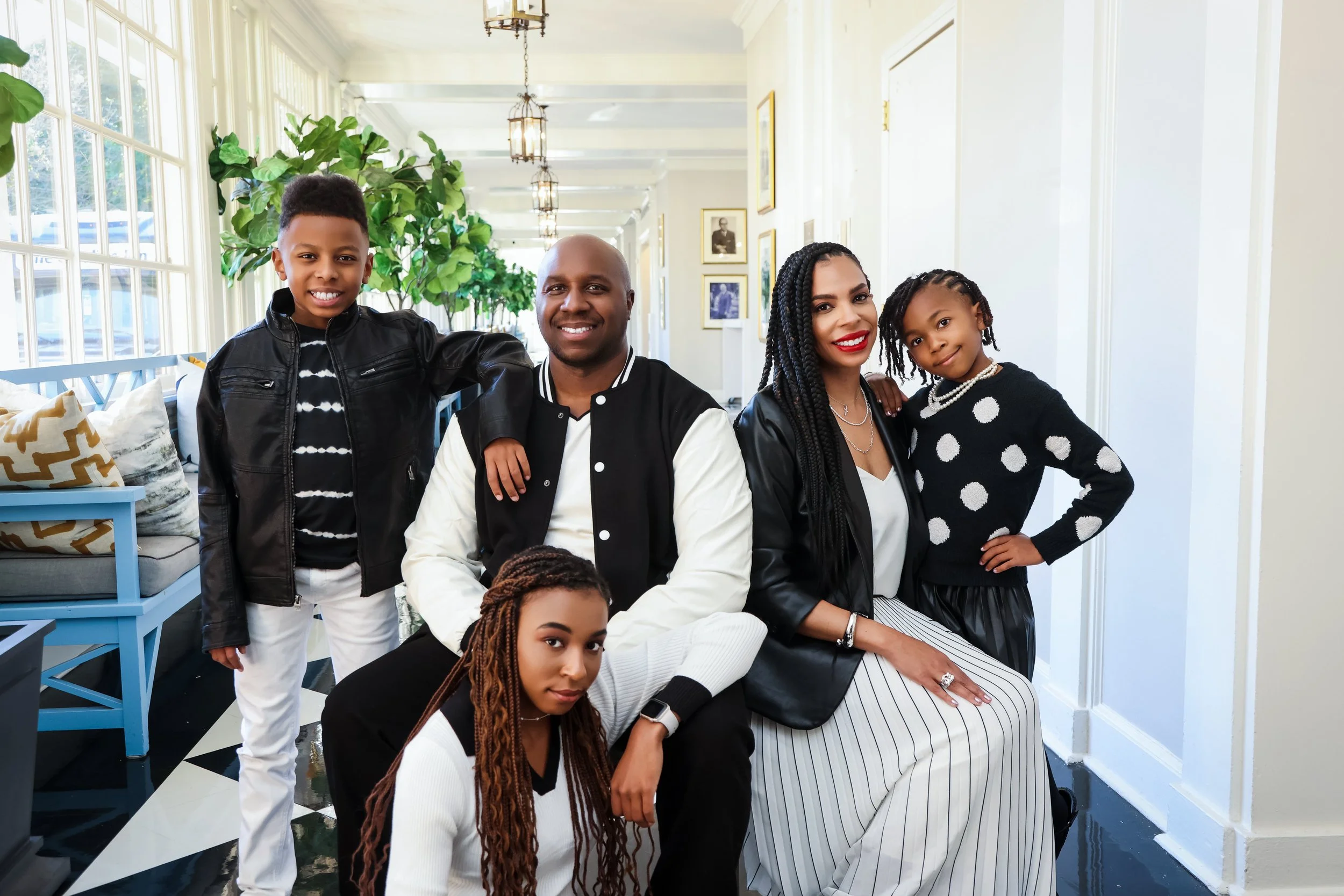 A family of five posing indoors, including a man, woman, two girls, and a boy. They are smiling and dressed in stylish clothing, with plants and framed pictures on the wall behind them.